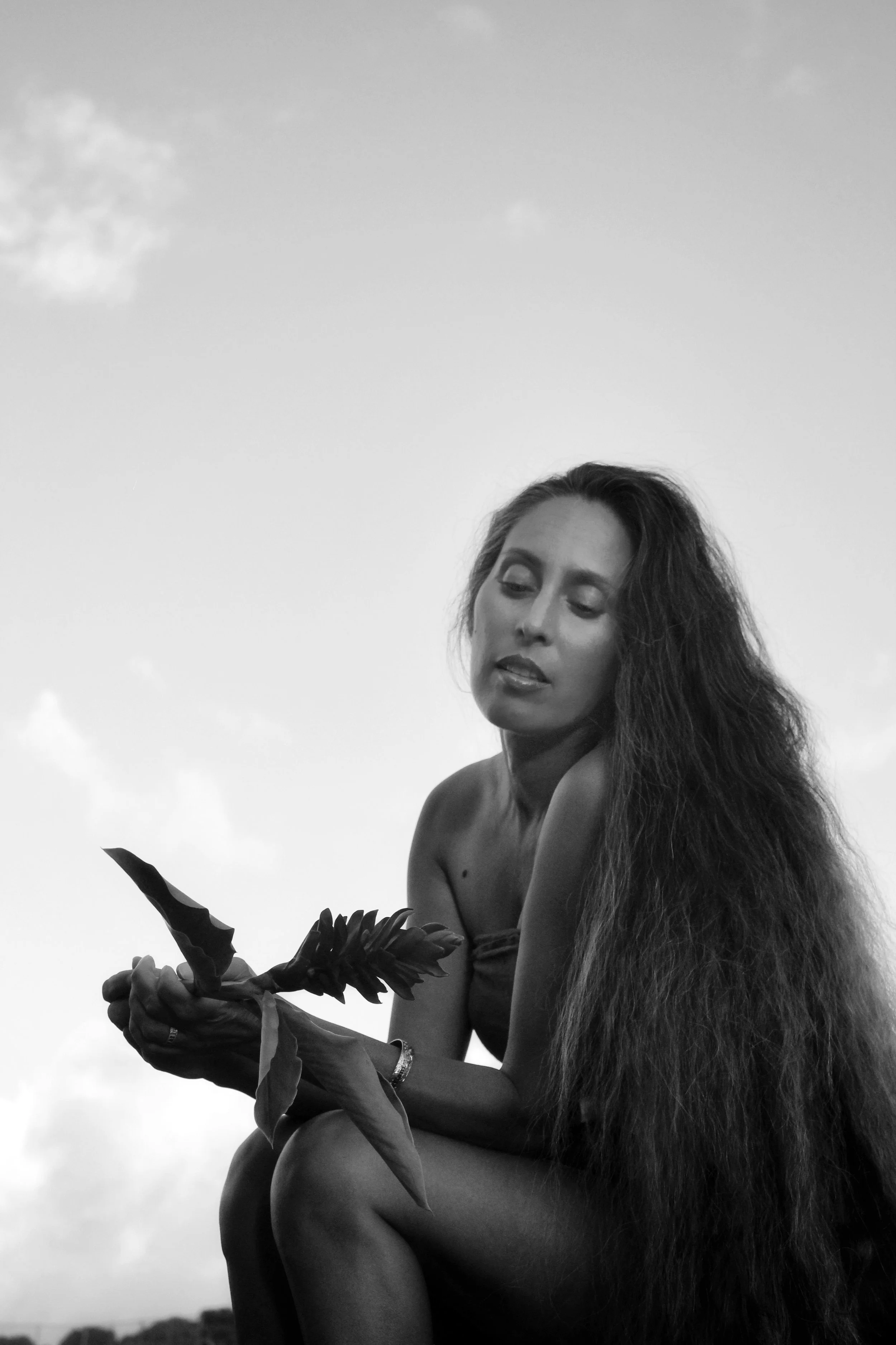 A black-and-white photo of a woman with long, curly hair sitting outdoors, holding a plant with broad leaves in her hands, against a cloudy sky background.