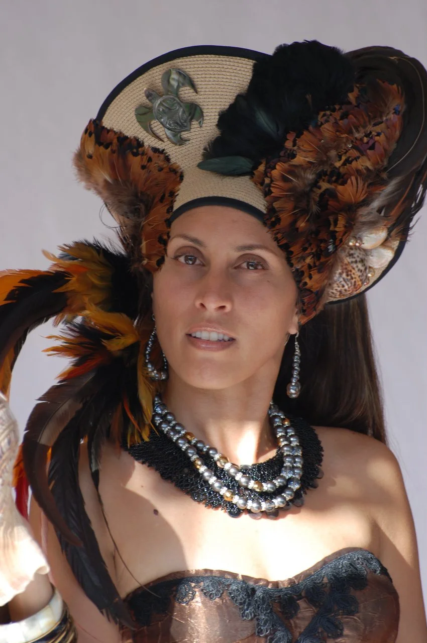 Woman wearing a large feathered hat adorned with a turtle pin, pearl earrings, layered necklaces, and a strapless dress with lace details.