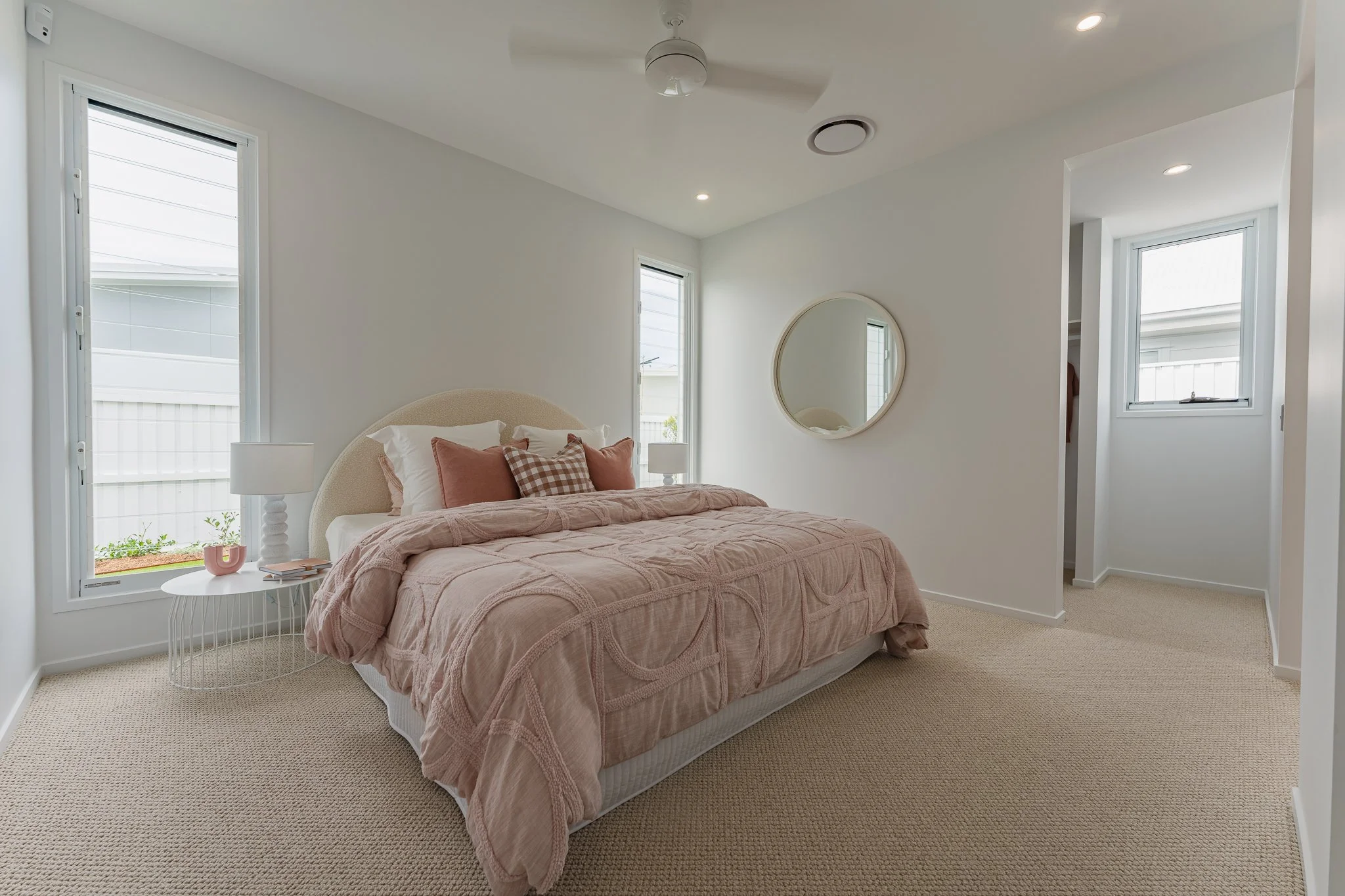 Bright bedroom with a large bed, pink bedding, and multiple pillows, two windows, a round mirror on the wall, and a side table with a lamp and decorative objects.