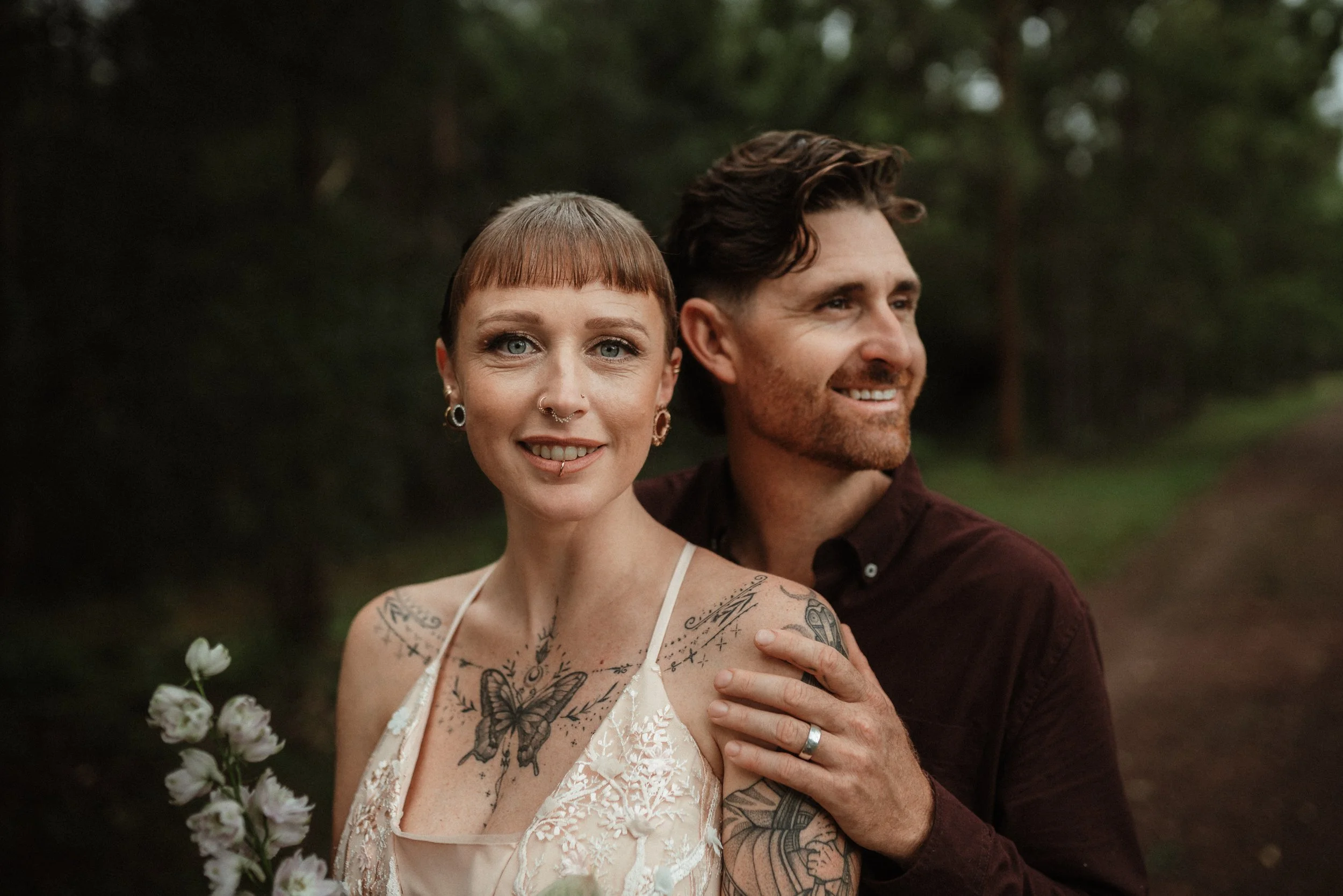 A couple outdoors, the woman holding a bouquet of flowers, both smiling, with trees in the background.