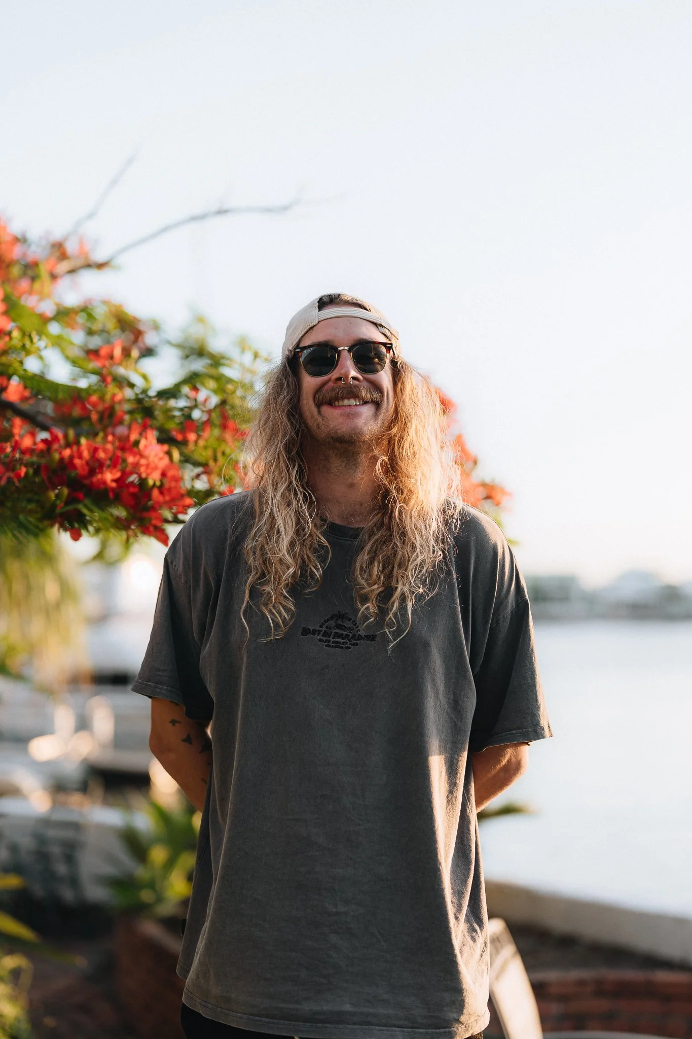 A man with long curly hair, wearing sunglasses, a backward baseball cap, and a dark t-shirt, standing outdoors near a body of water with blooming trees in the background.