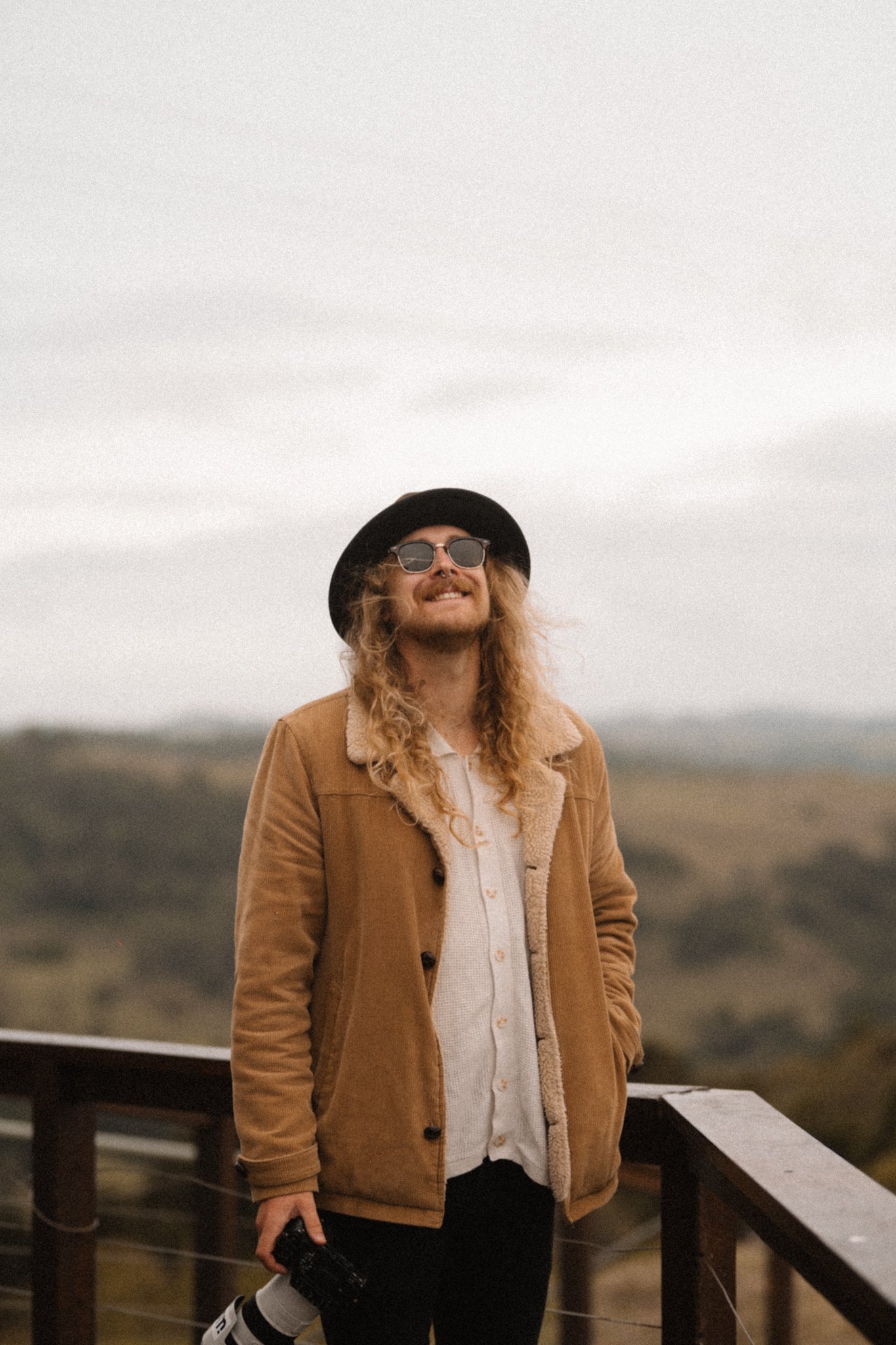 A man with long curly hair, wearing sunglasses, a black hat, a brown jacket with a shear lining, and a white shirt, standing outdoors on a cloudy day on a wooden platform.