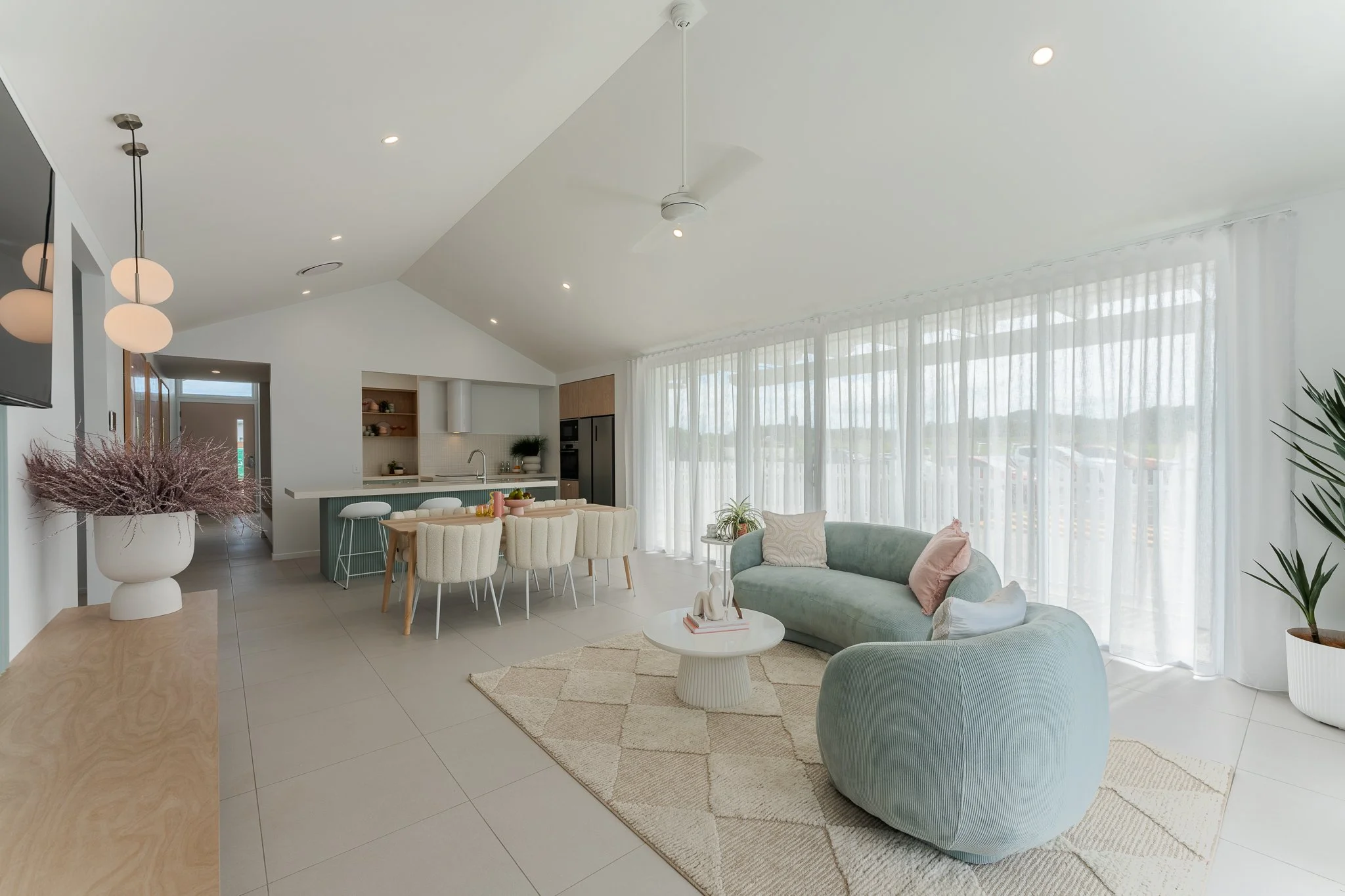 Bright, modern living room with pastel-colored furniture, large windows with sheer curtains, a dining area, and a kitchen in the background.