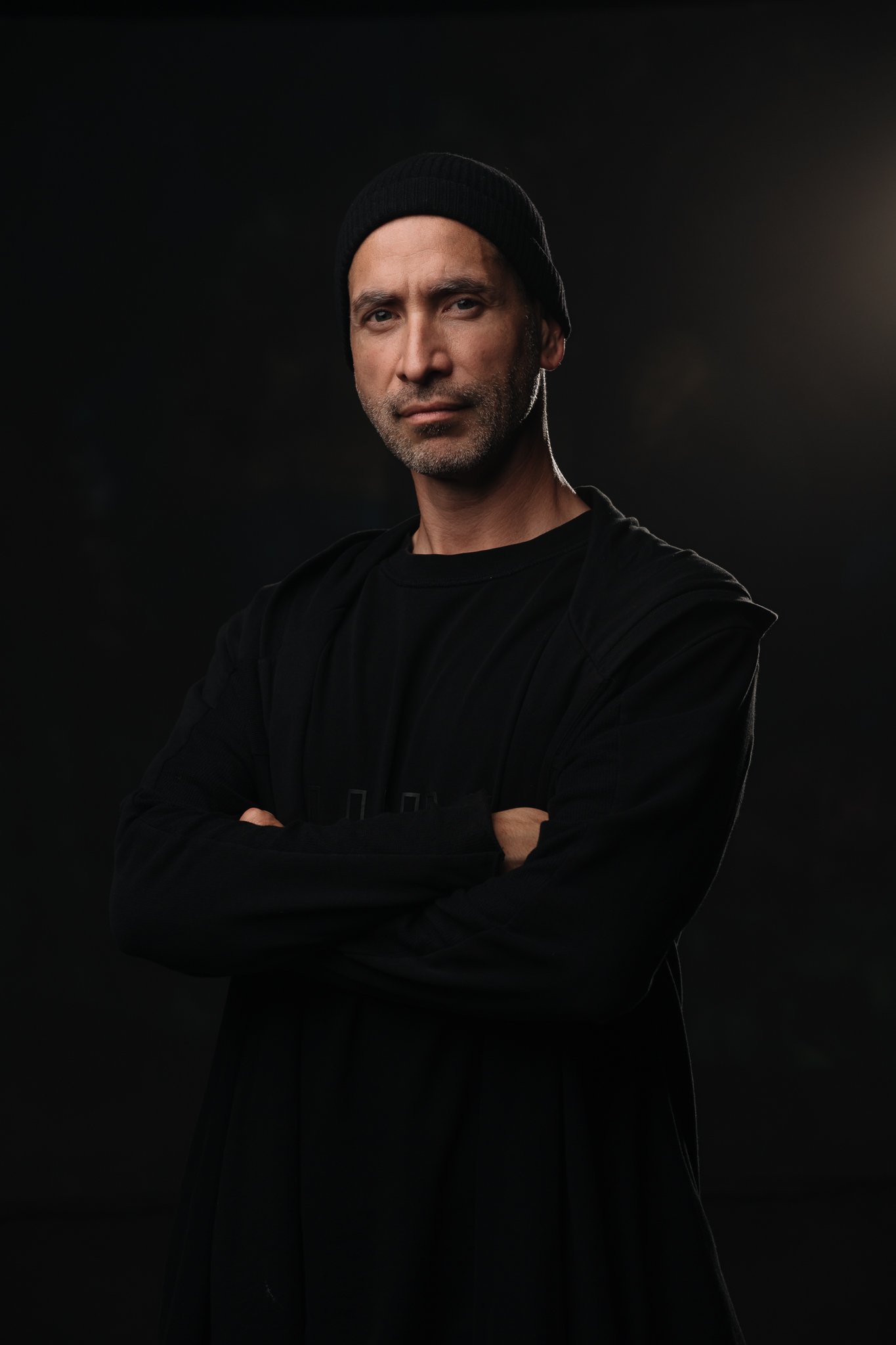 A man wearing a black beanie and black clothing stands with arms crossed against a dark background, illuminated by a light source from the right.