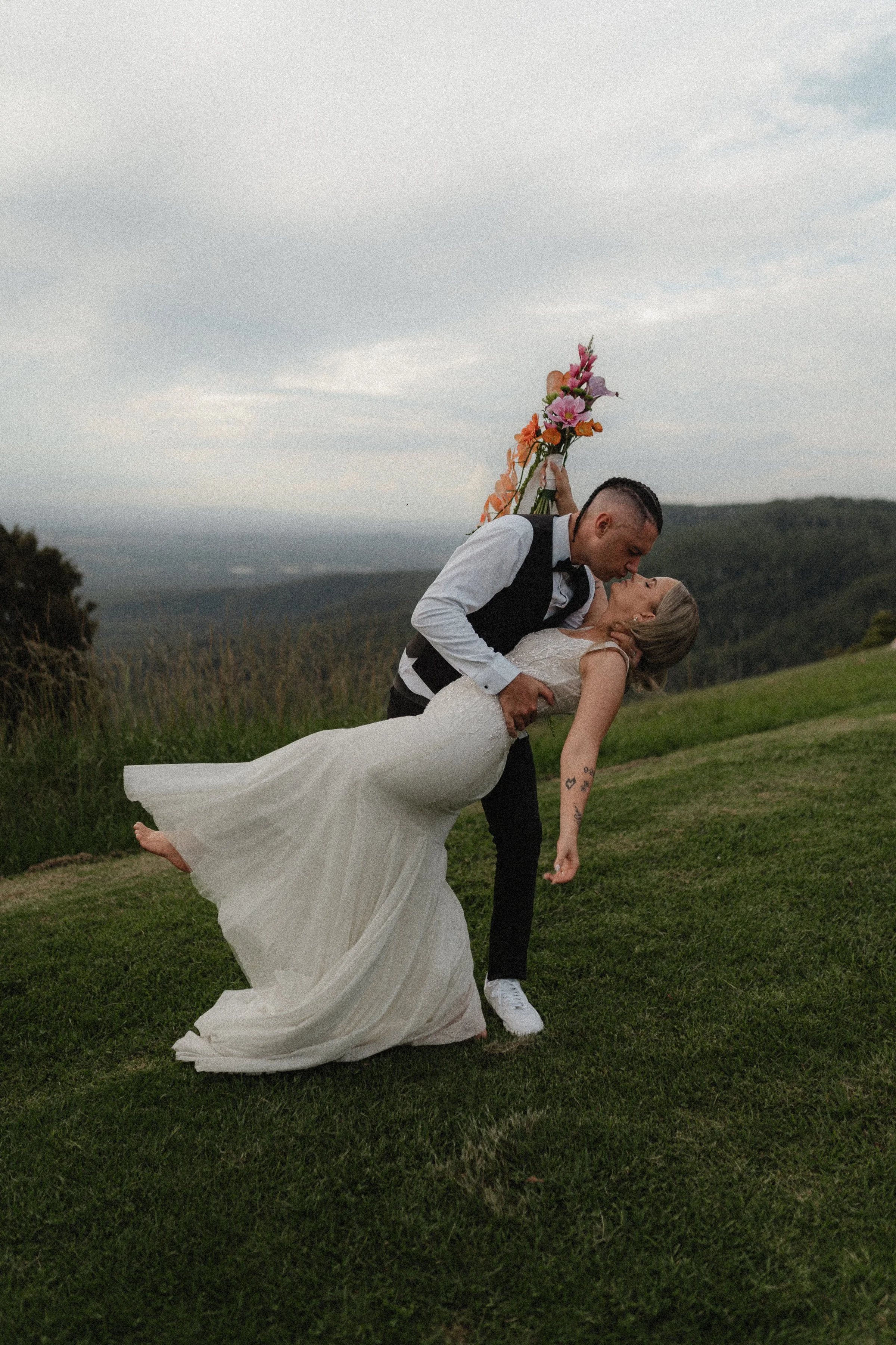 A groom in a tuxedo and a bride in a white wedding dress sharing a kiss outdoors on a grassy hill with mountains in the background, the groom holding a bouquet of colorful flowers.