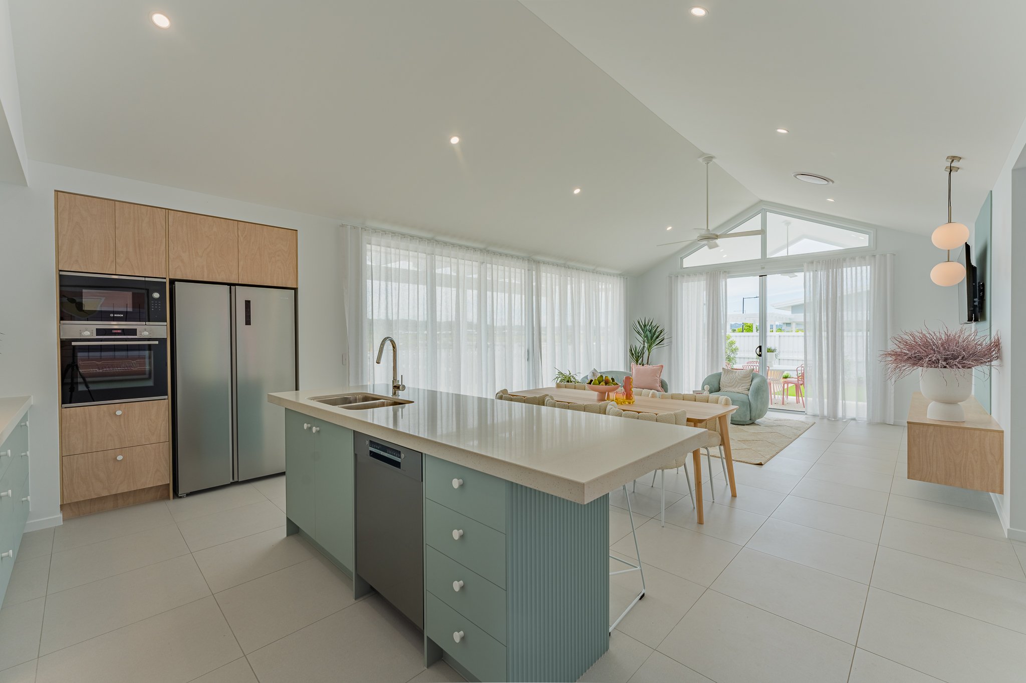 Open-concept modern kitchen and living room with large windows, white curtains, and minimalist decor, including pastel-colored furniture and potted plants.
