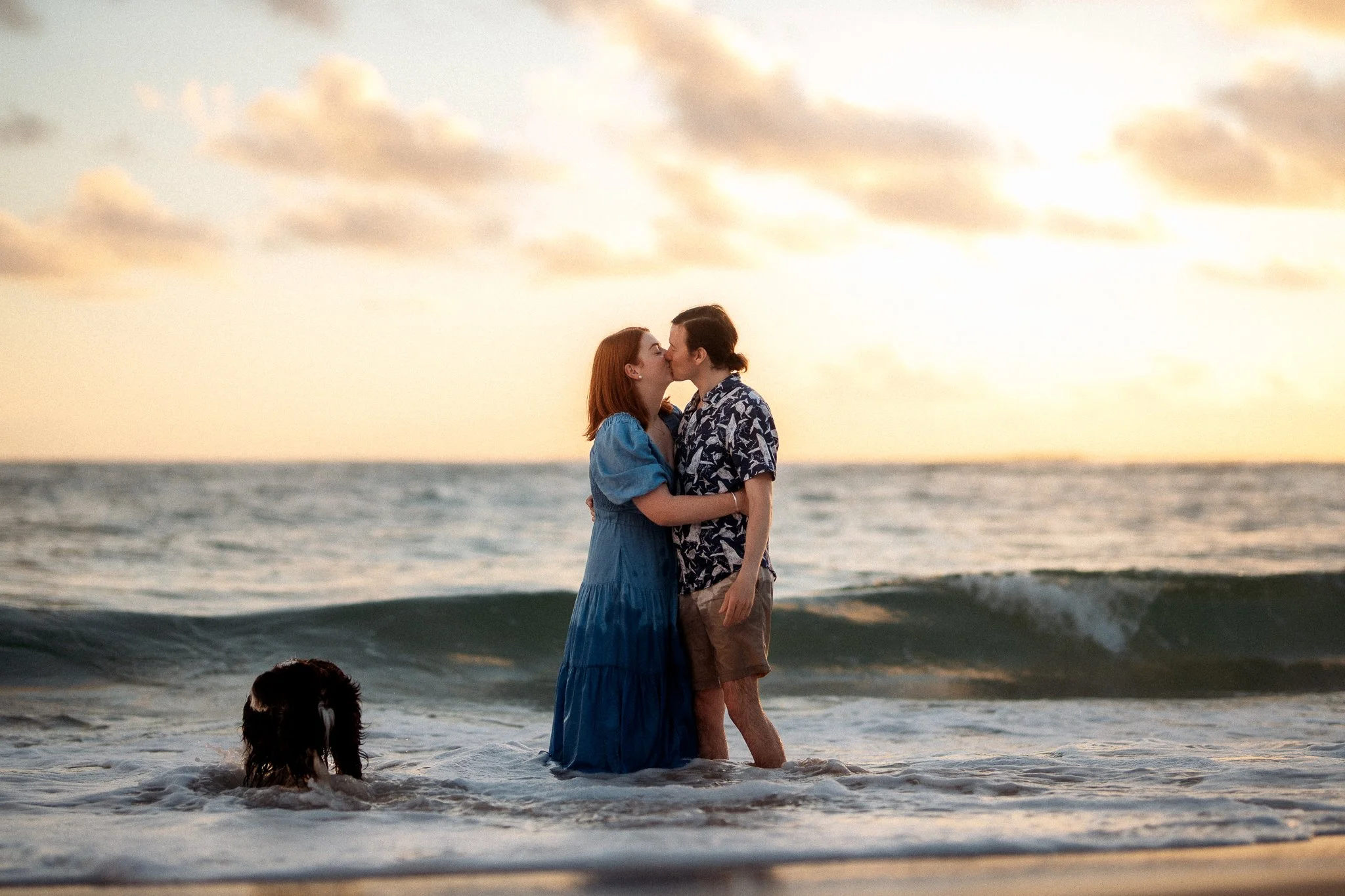 A couple in wedding attire kissing in the ocean at sunset, with a dog swimming nearby.