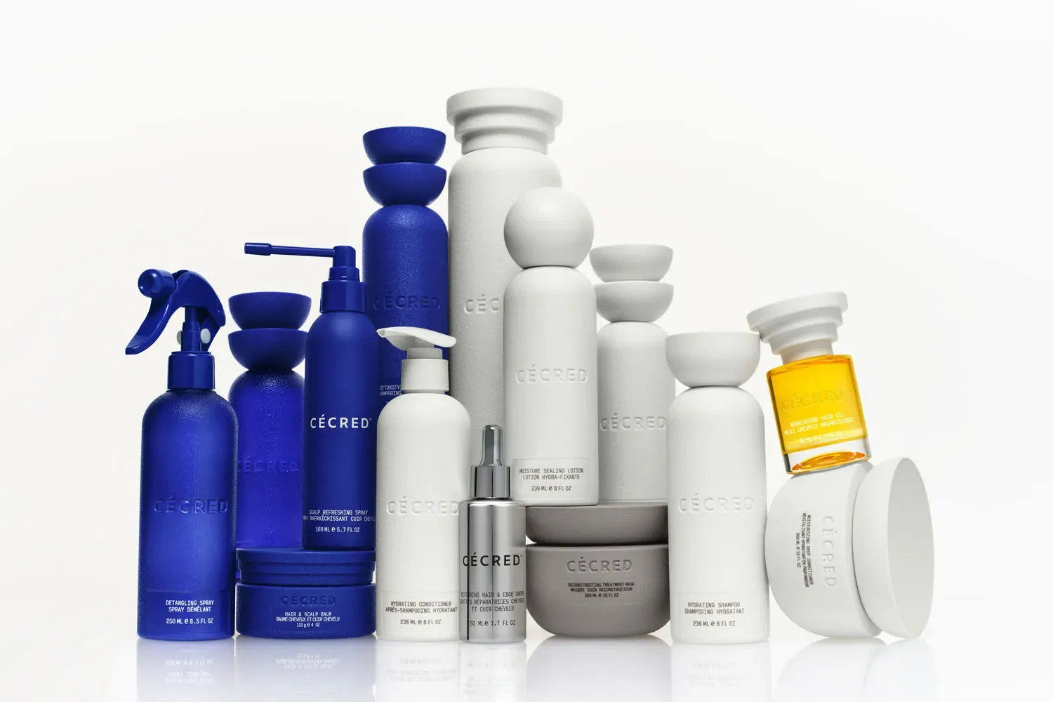 A collection of CÉCRED skincare products arranged on a white surface, including bottles, jars, and tubes in blue, white, yellow, and silver packaging.