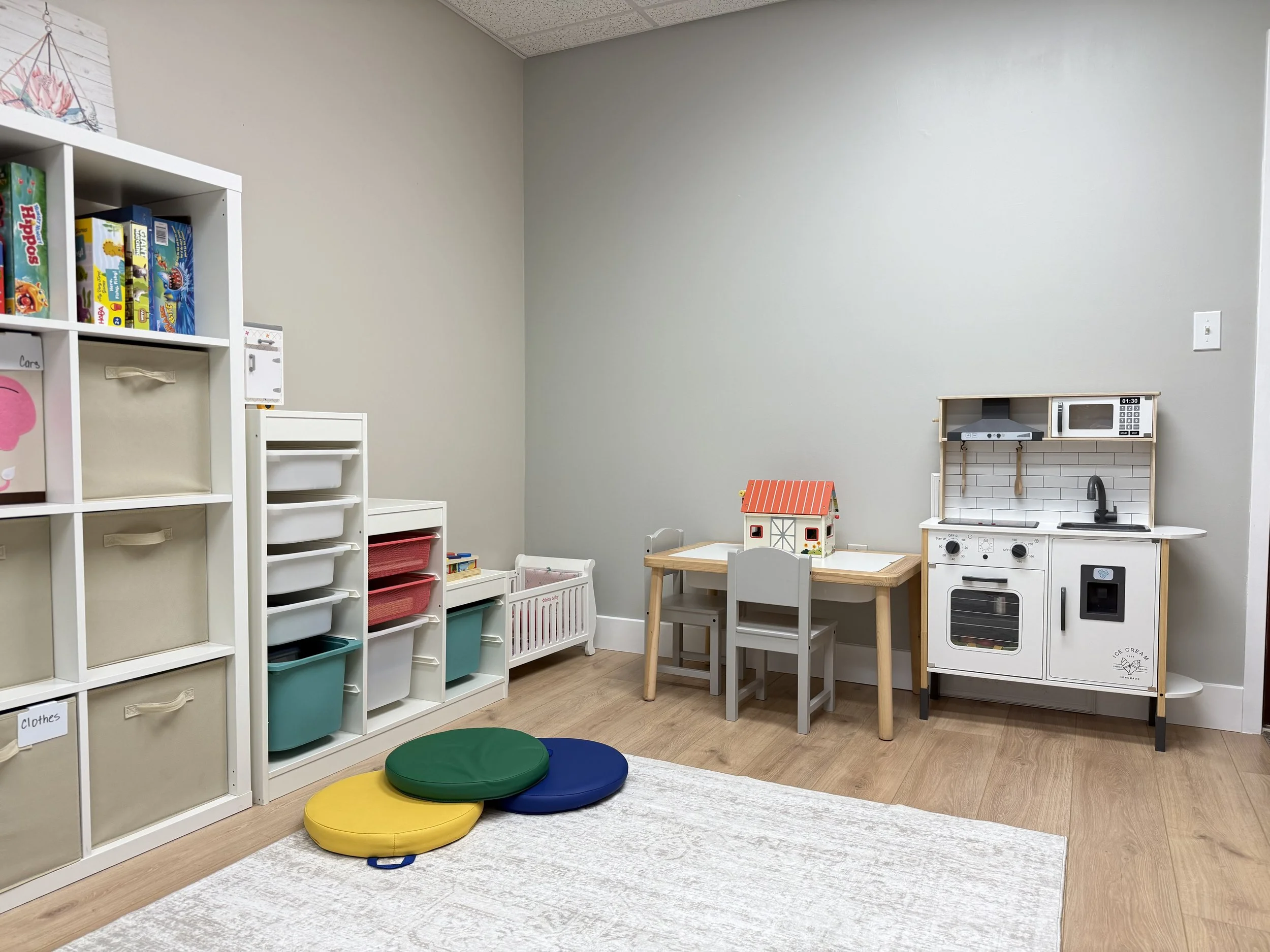 Playroom with toy kitchen set, small table, chairs, storage bins, and colored floor cushions.