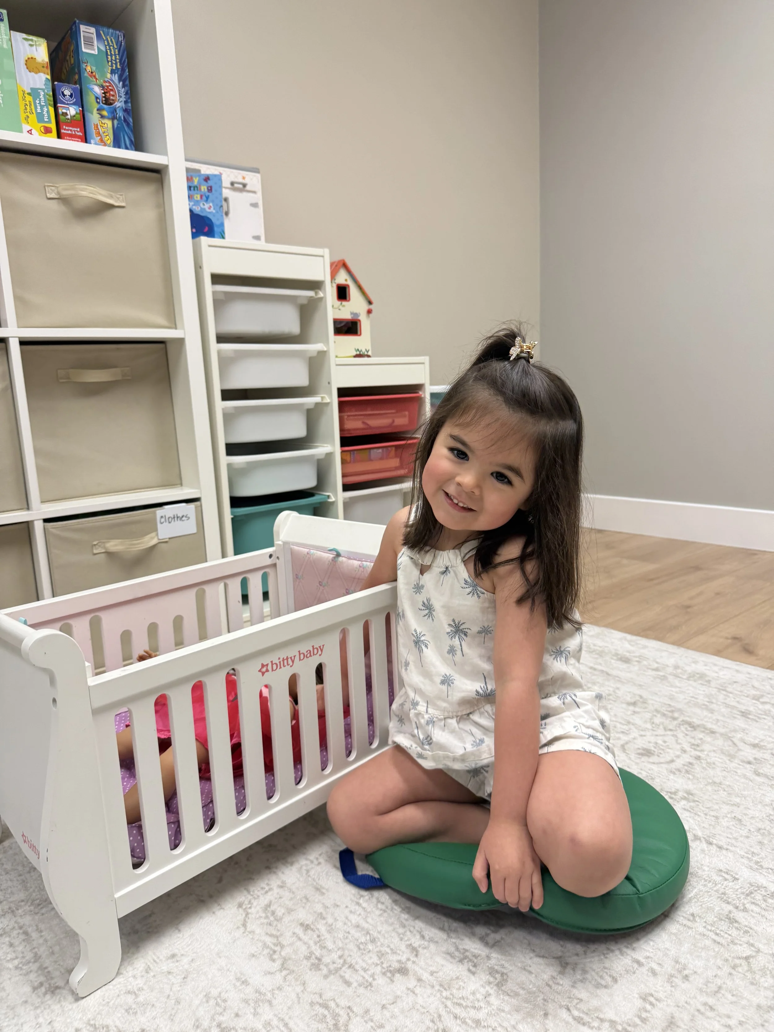 A young girl with dark hair, wearing a sleeveless dress with palm tree prints, sitting on a green cushion next to a toy crib with a doll inside, in a room with storage shelves and a beige rug.