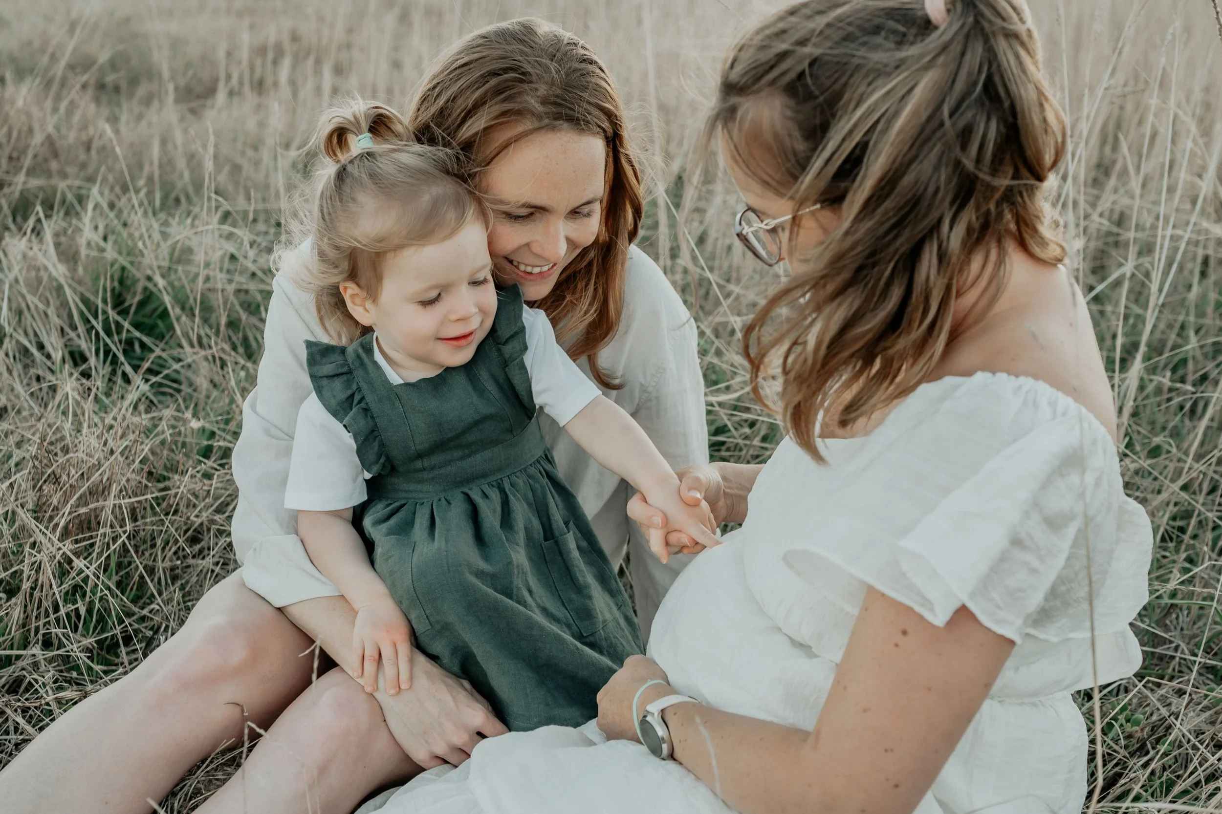 A little girl sitting with her two mothers pointing at her mother's pregnant belly