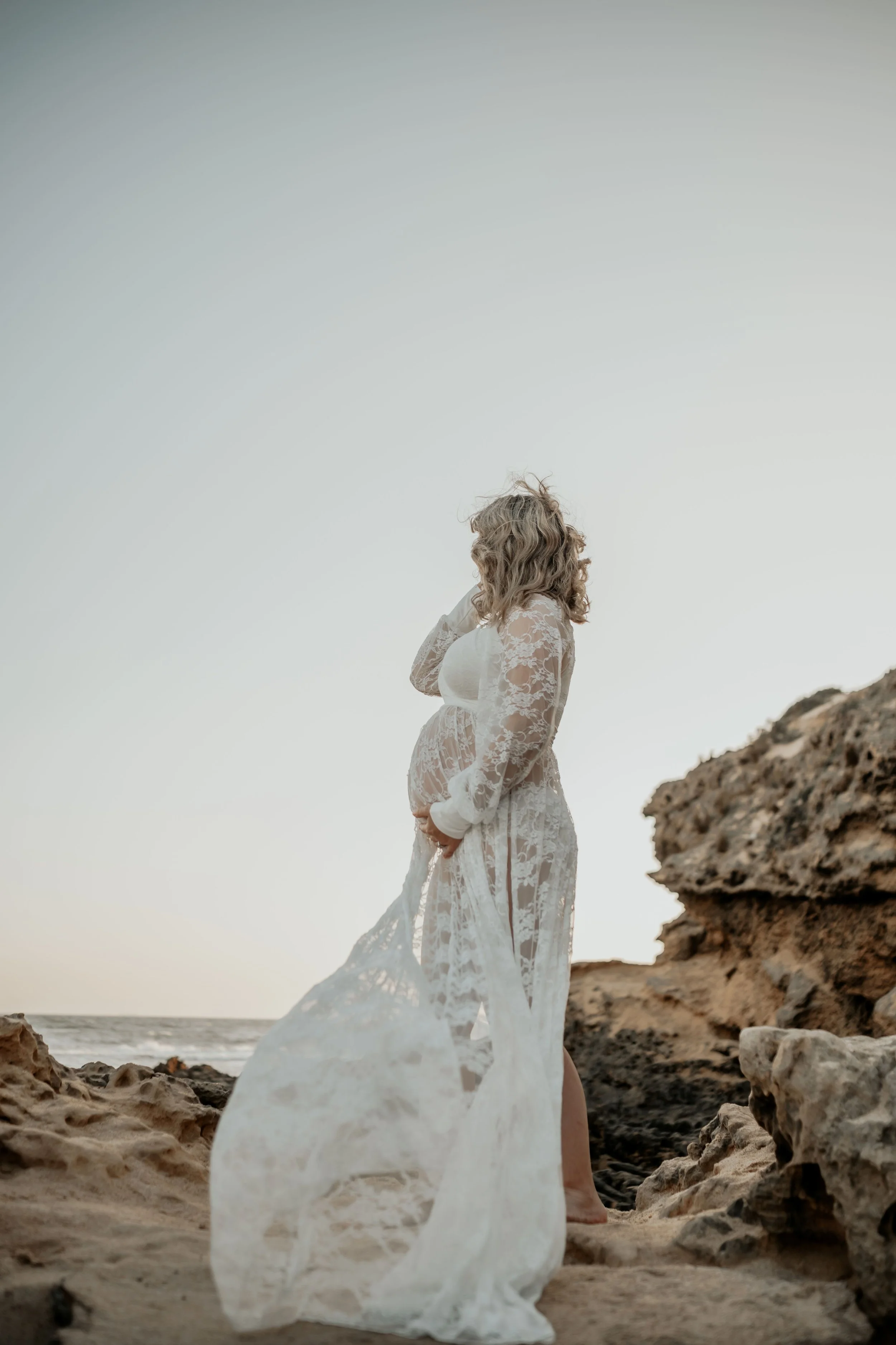 A pregnant woman looks out at the wild ocean while holding her belly and hair