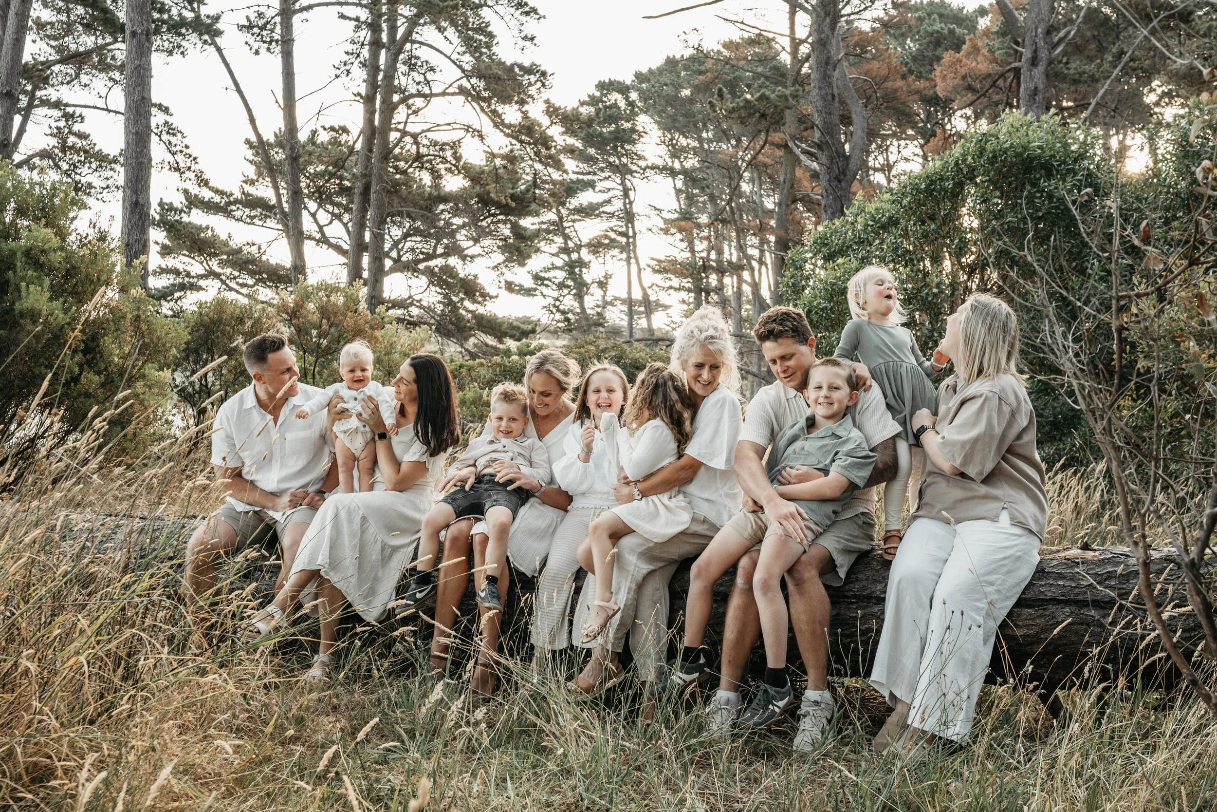 A family group of nine people, including children, sitting on a log in a forest setting. The adults wear light-colored clothing, and the children are in casual outfits. Trees and grass surround them under a bright sky.