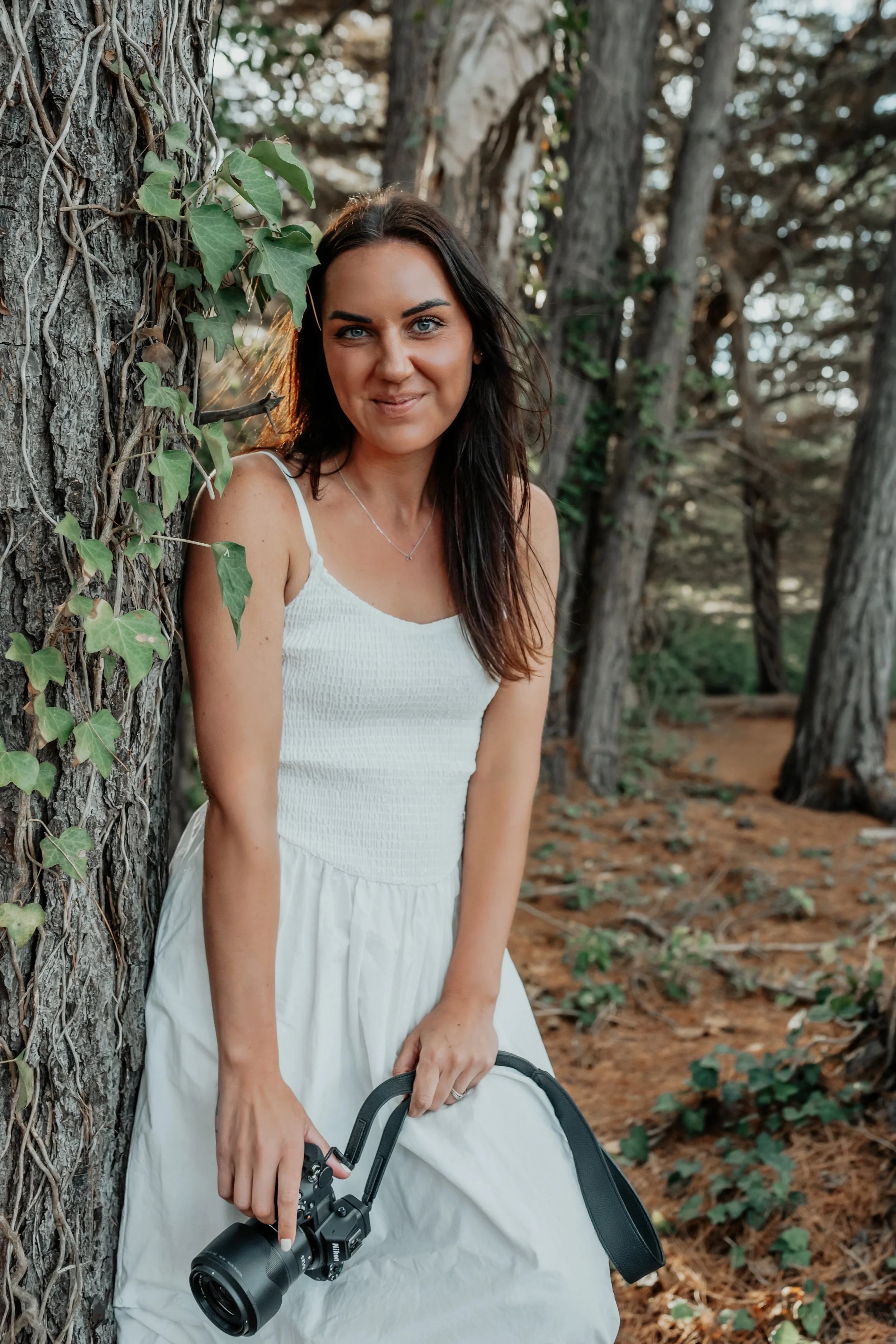 Bek standing against a tree grinning and holding her camera