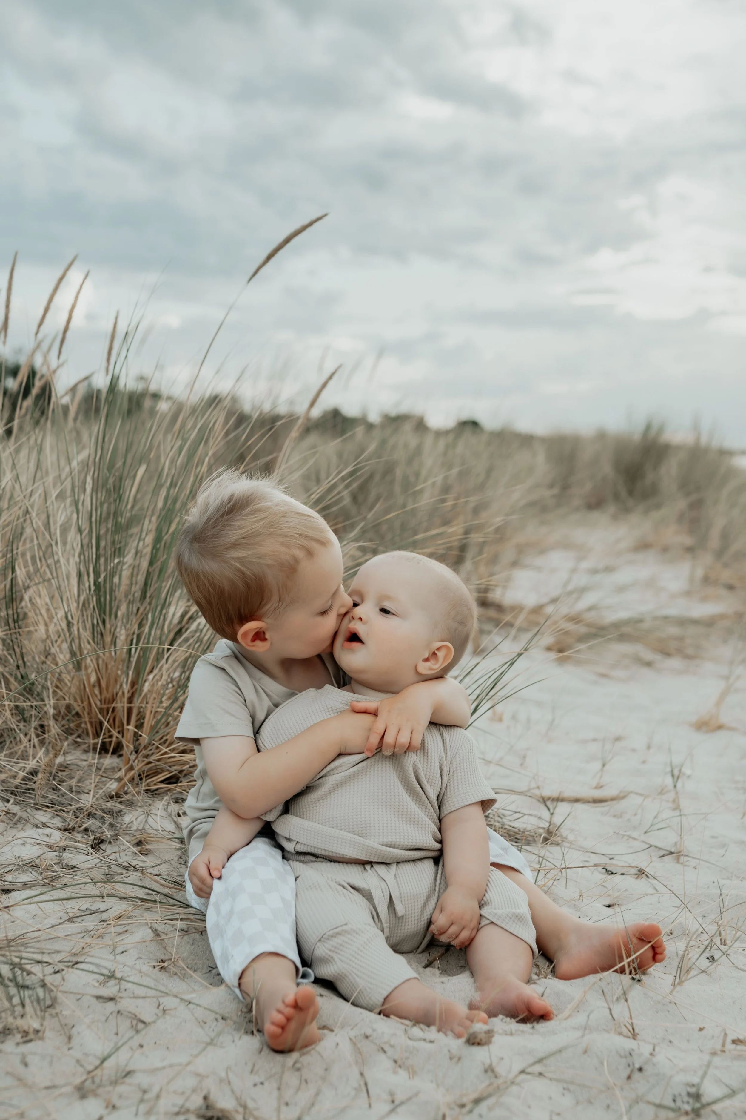 Two little brothers sitting on the sand. Big brother giving little brother a kiss on the cheek