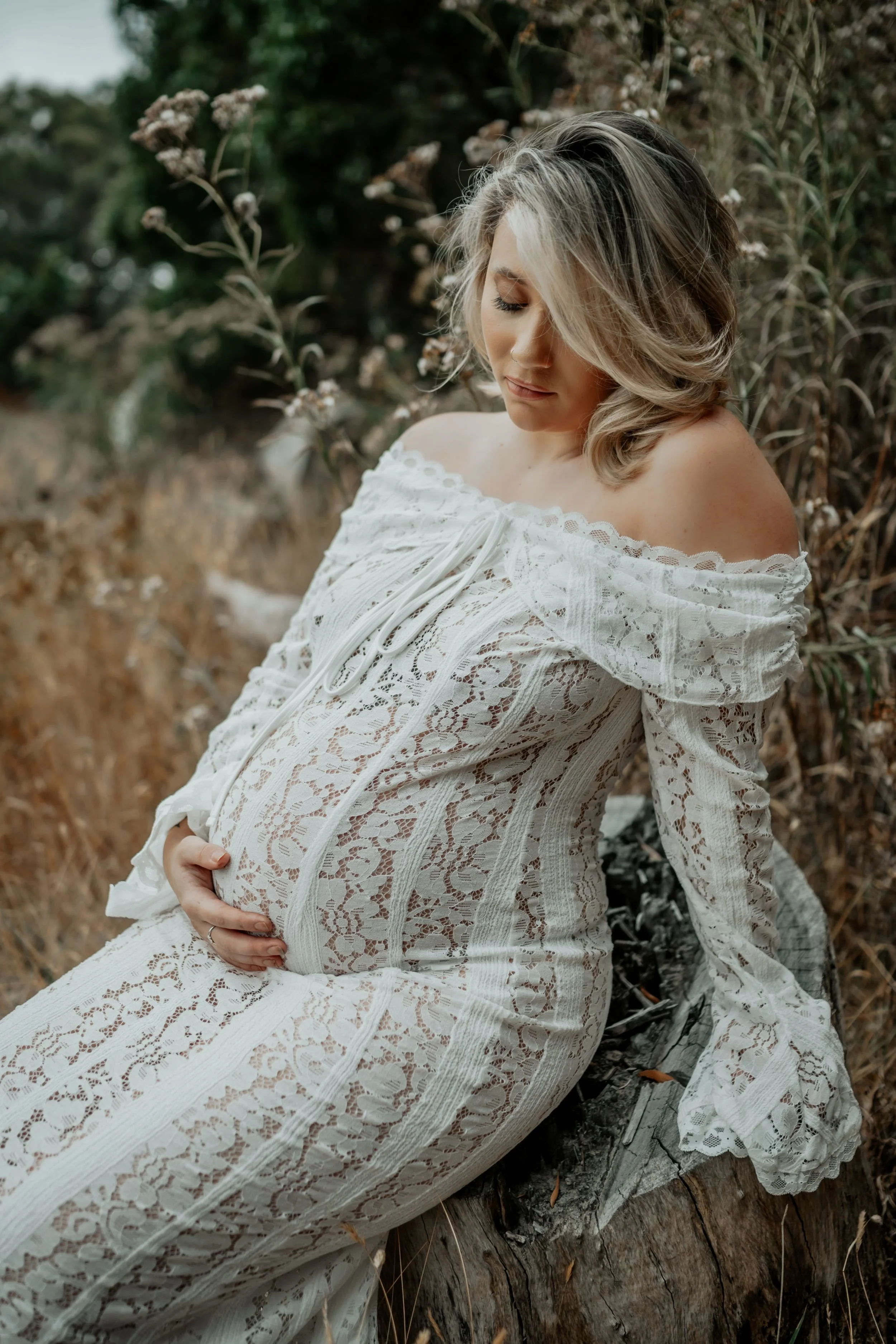 A pregnant woman sits on a log in the bush looking down at her unborn baby