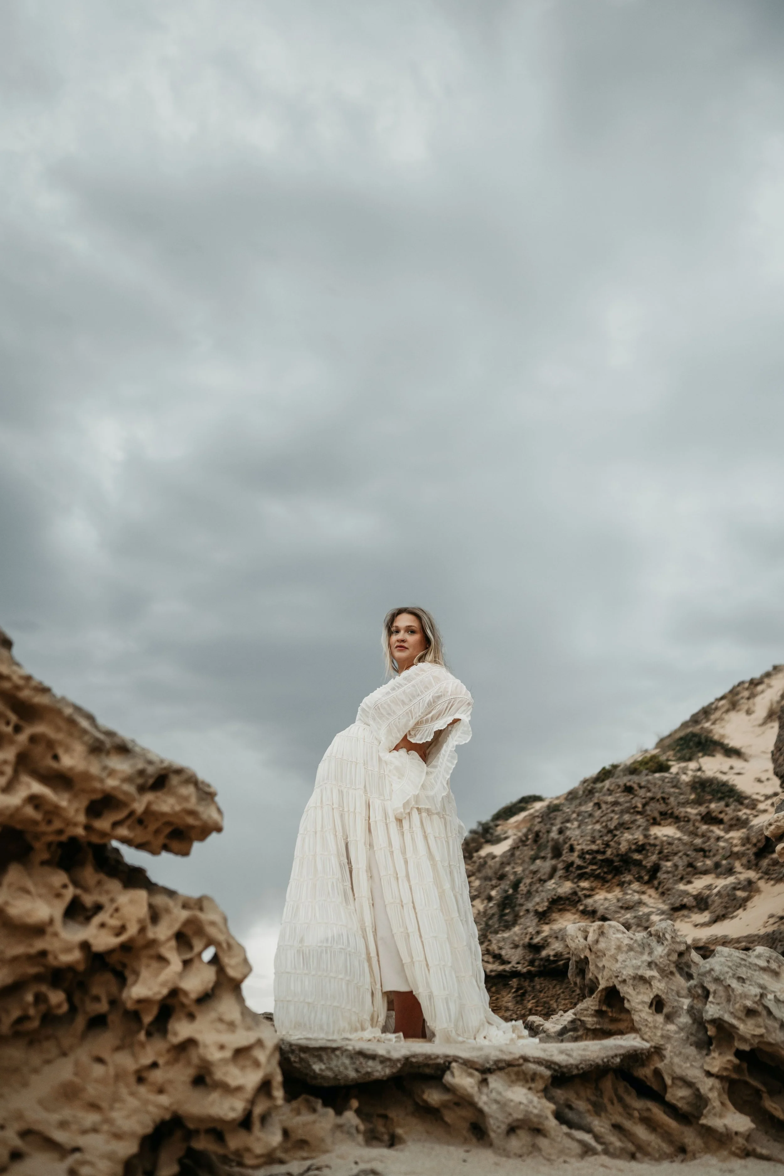 A pregnant woman stands on a rock formation in a gown