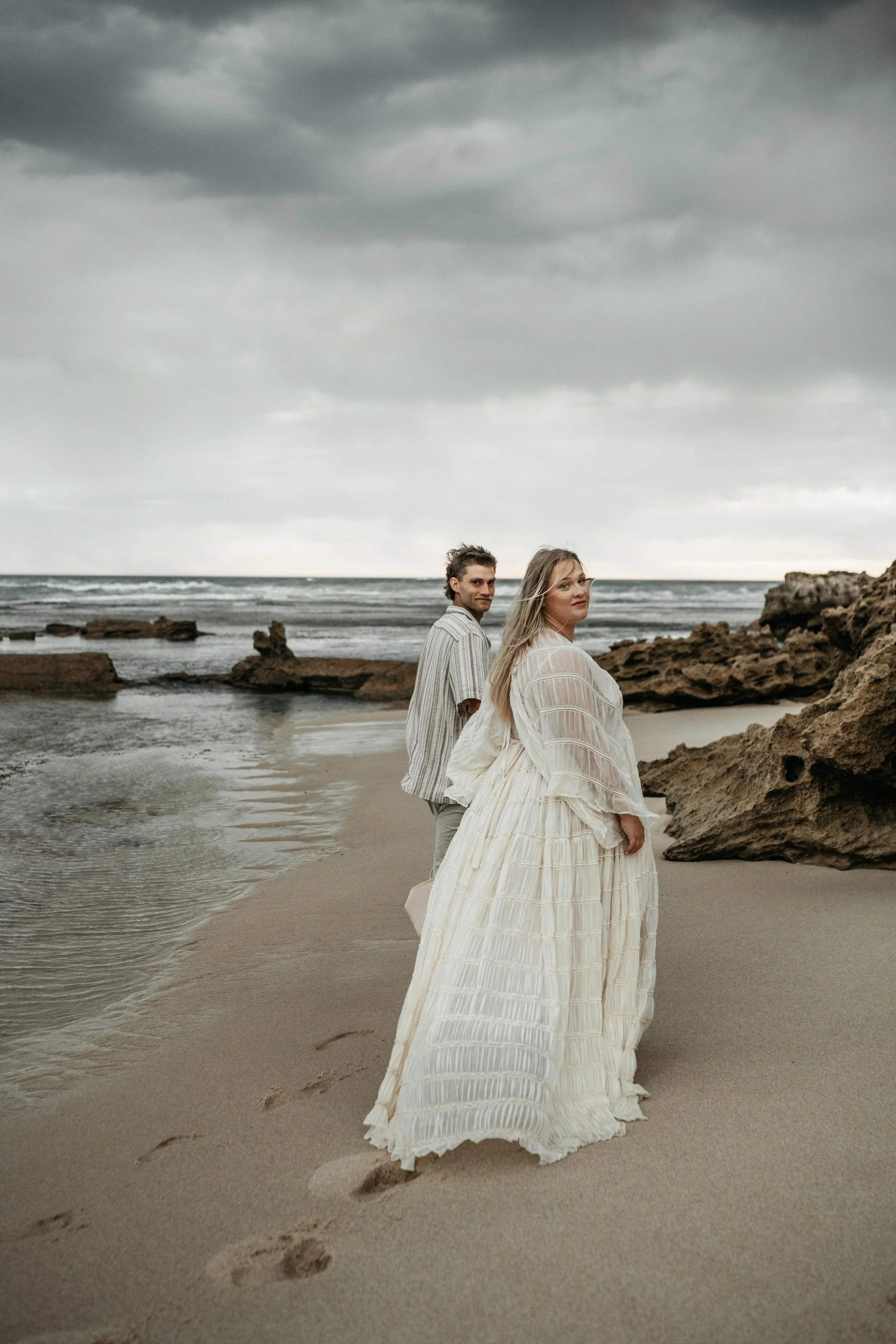 A pregnant couple walking along the beach looking at the camera as they walk away holding hands