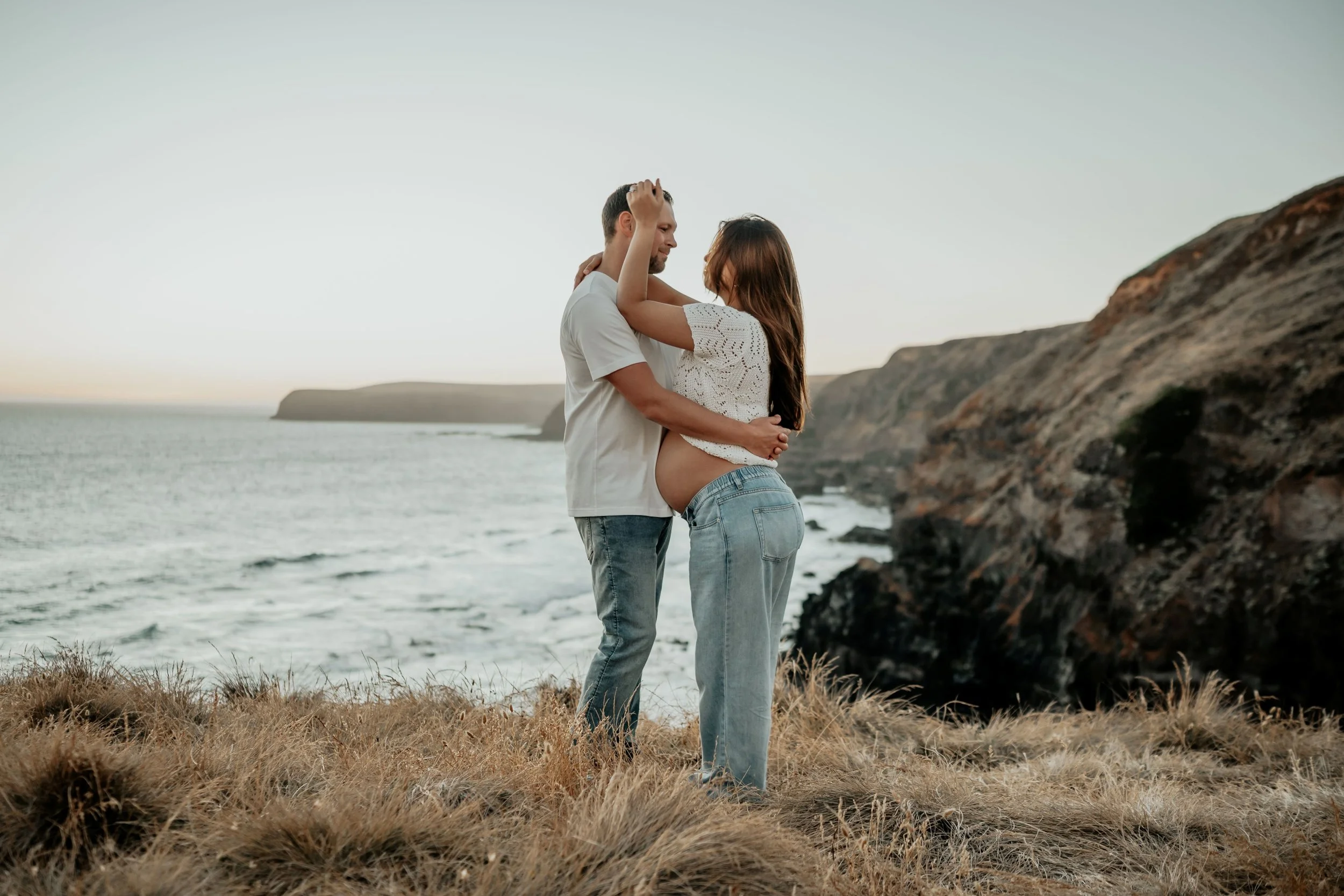 A pregnant women plays with her husbands hair with a stunning backdrop of a cliff hill and open ocean