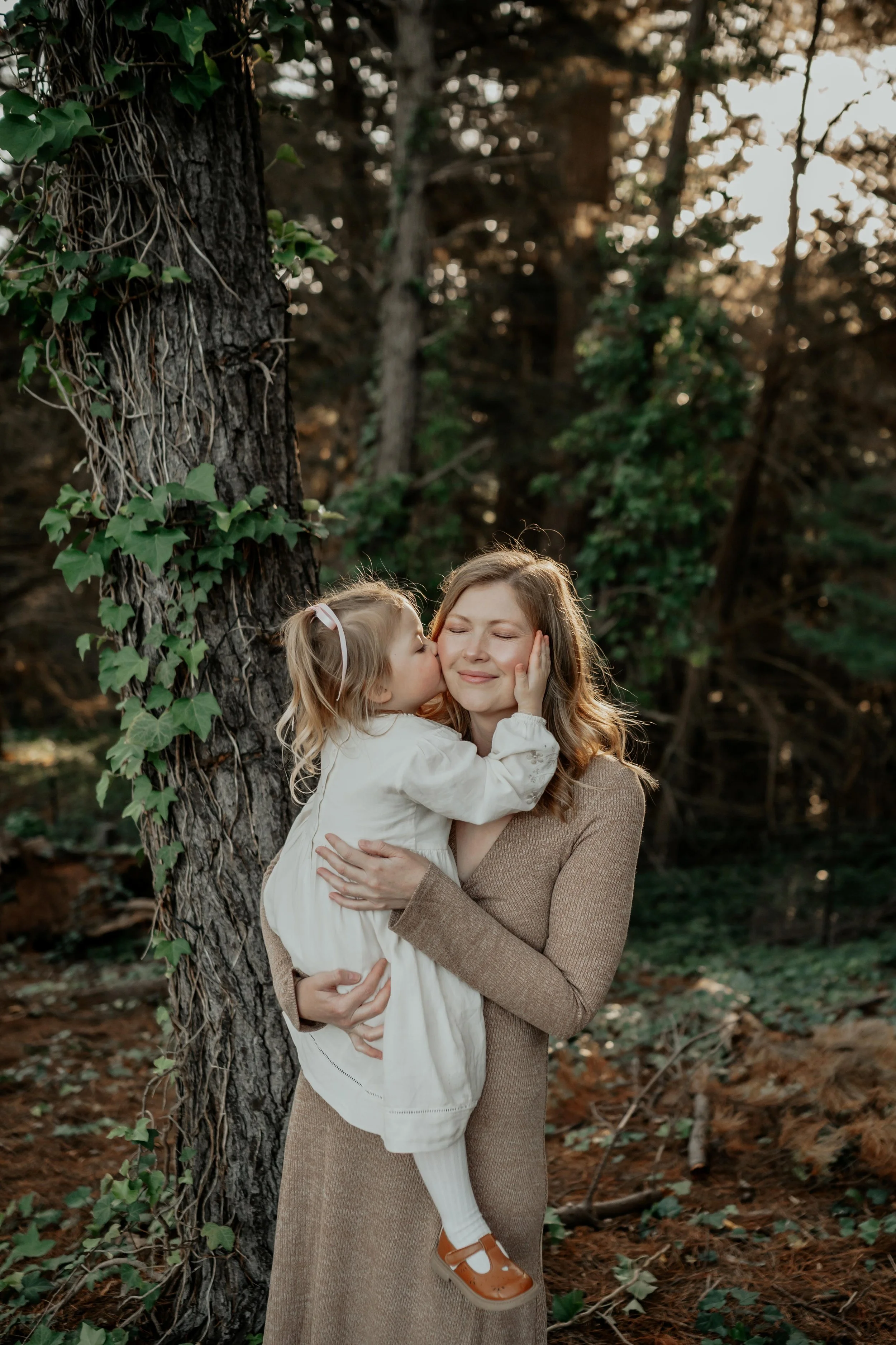 A mother standing in the forest holding her daughter. Daughter holding her mum's face with her hand and kissing her cheek