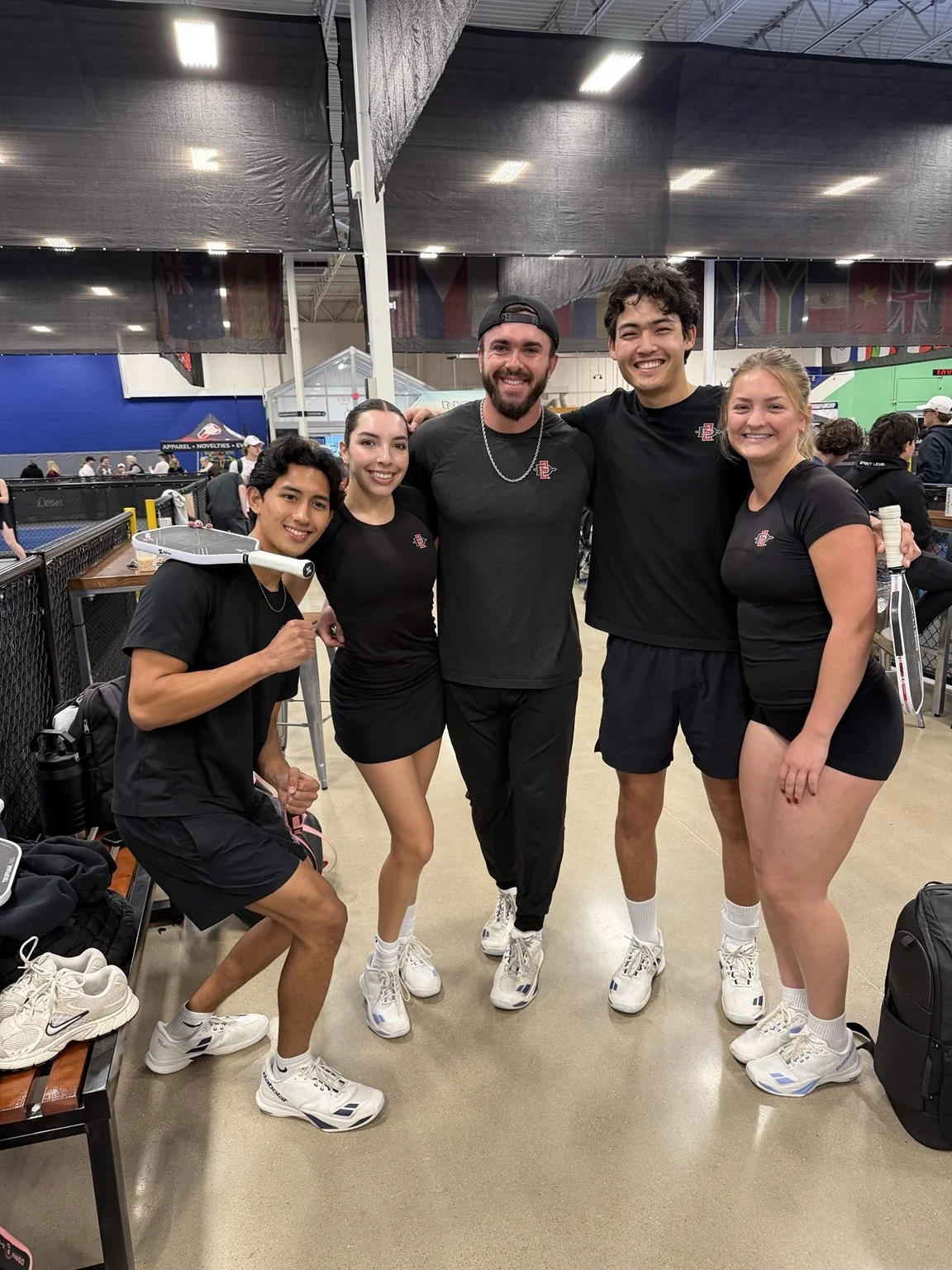Arden Light and Elitza Lopez with fellow players beside an indoor pickleball court.