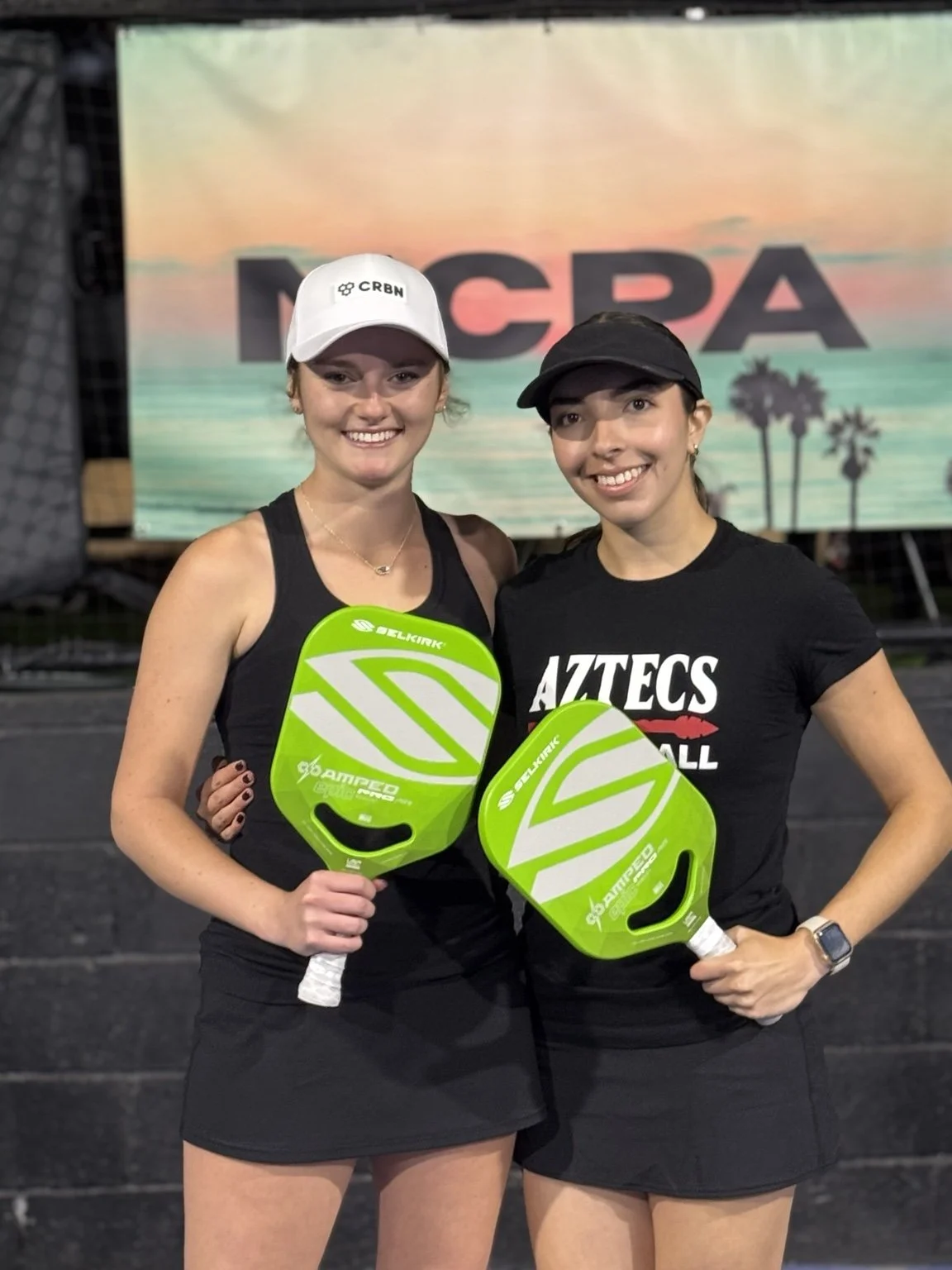 Elitza Lopez and a fellow player smiling with green pickleball paddles at an NCPA event.