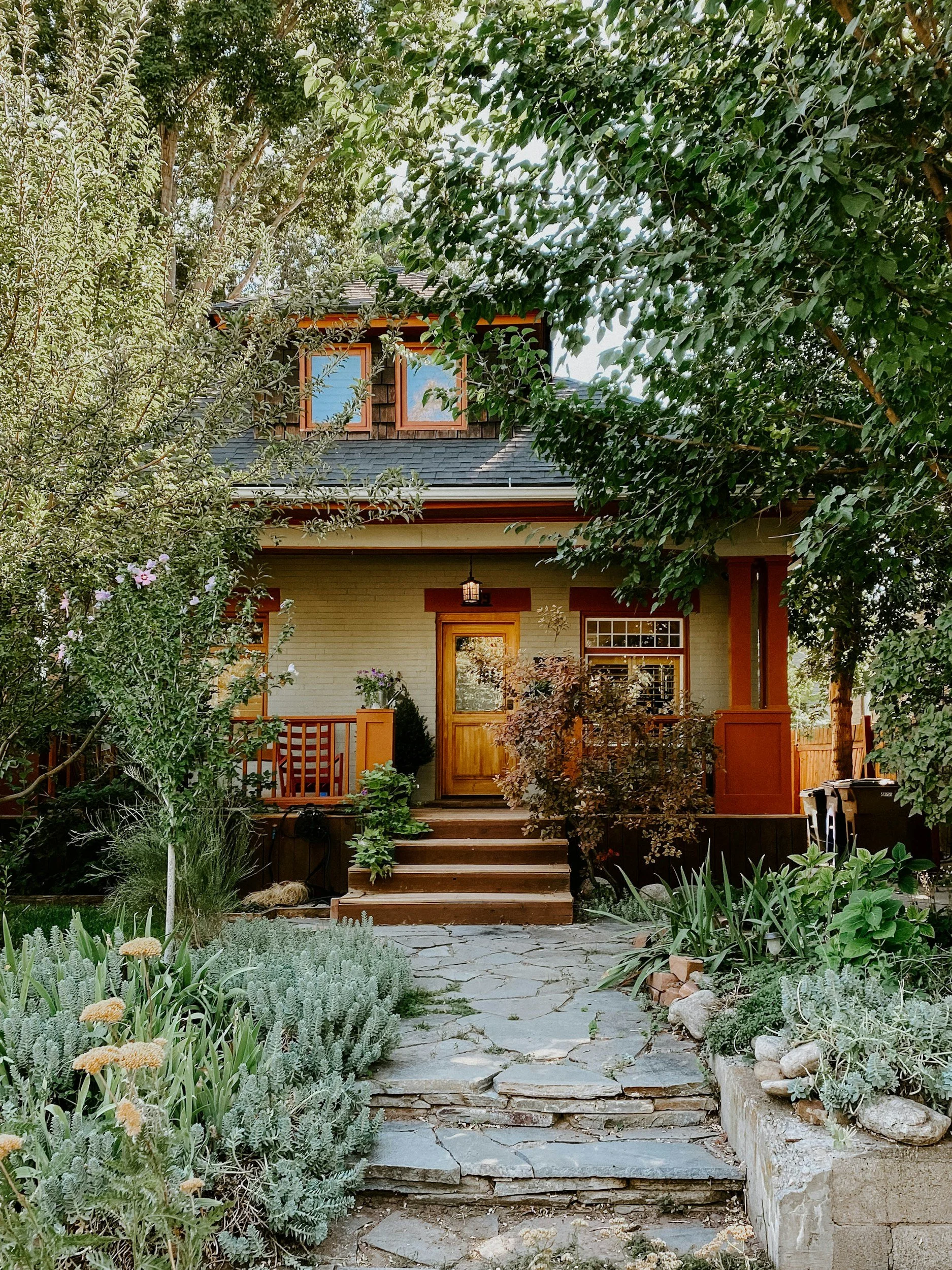 Front yard of a house with stone pathway, plants, trees, and a wooden porch with steps leading to the front door