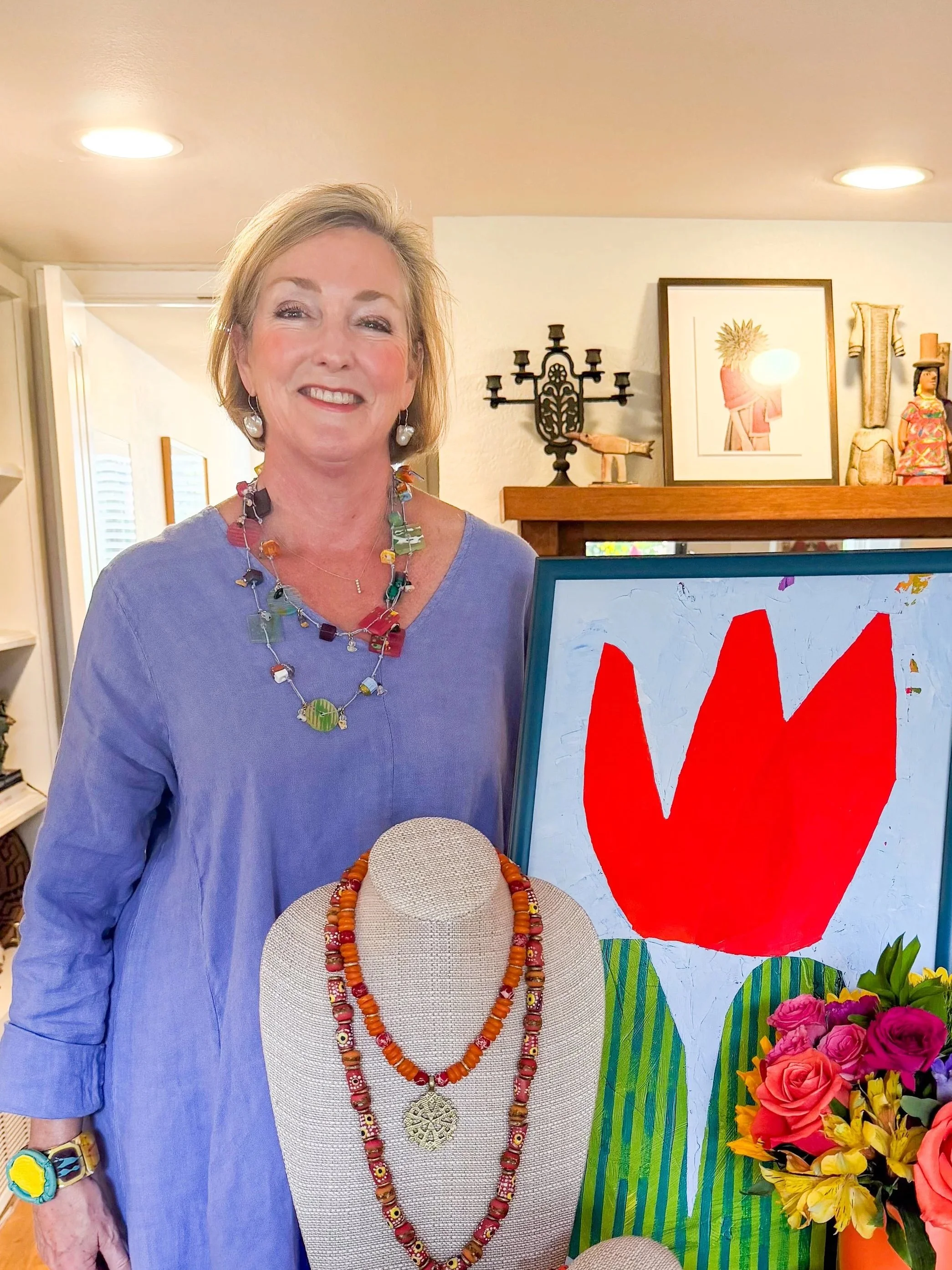 A smiling woman with blonde hair wearing a purple top, colorful jewelry, and a floral bracelet displayed in front of a colorful art piece and a bouquet of pink, yellow, and orange flowers.