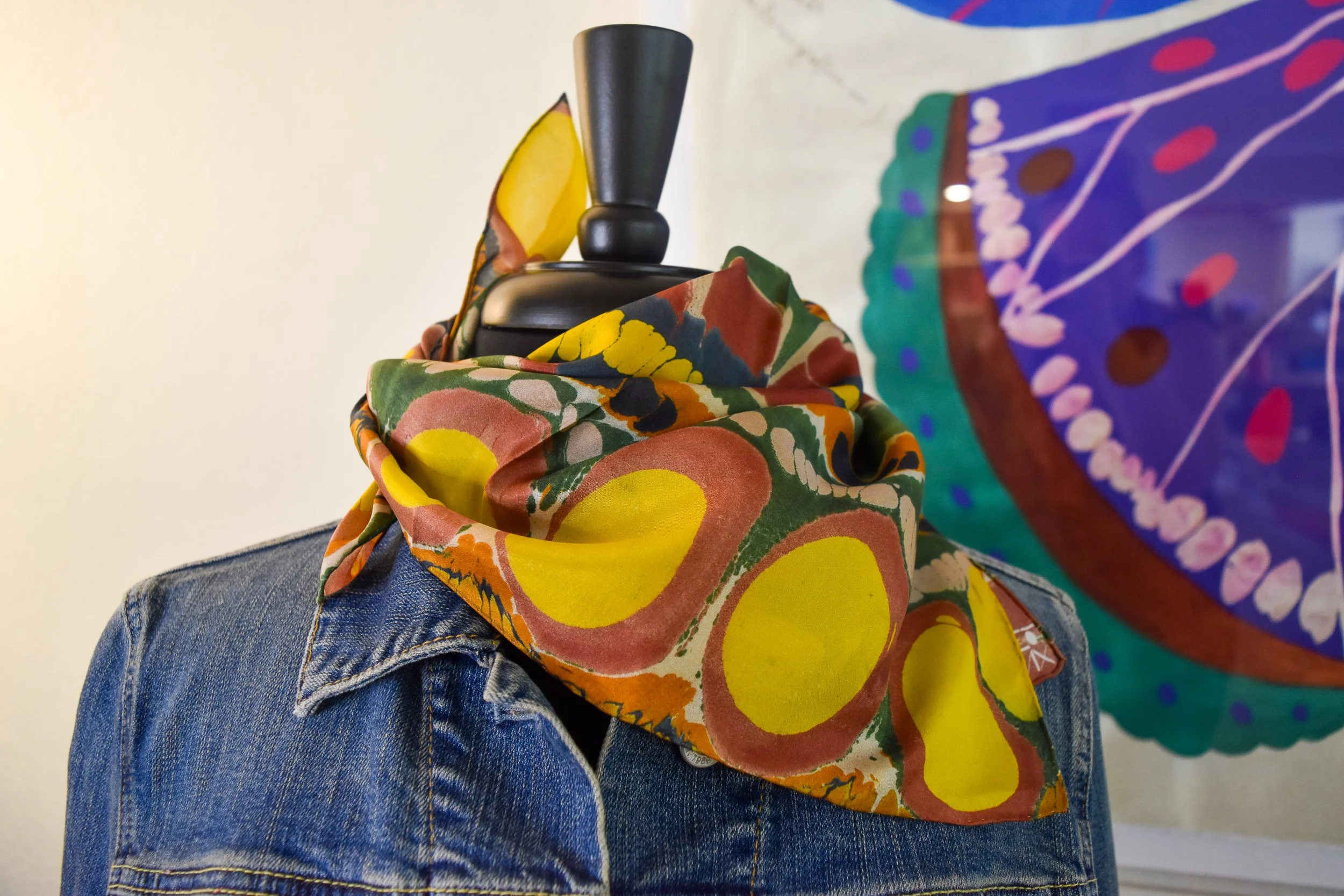 Close-up of a person wearing blue jeans, with a colorful patterned scarf or fabric draped around their neck. The background is a plain off-white wall.