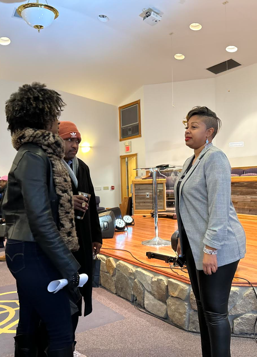Three people in conversation indoors near a stage with a stone wall and podium. One is holding a drink and wearing a red beanie, another with curly hair and a scarf, and the third in a gray blazer.
