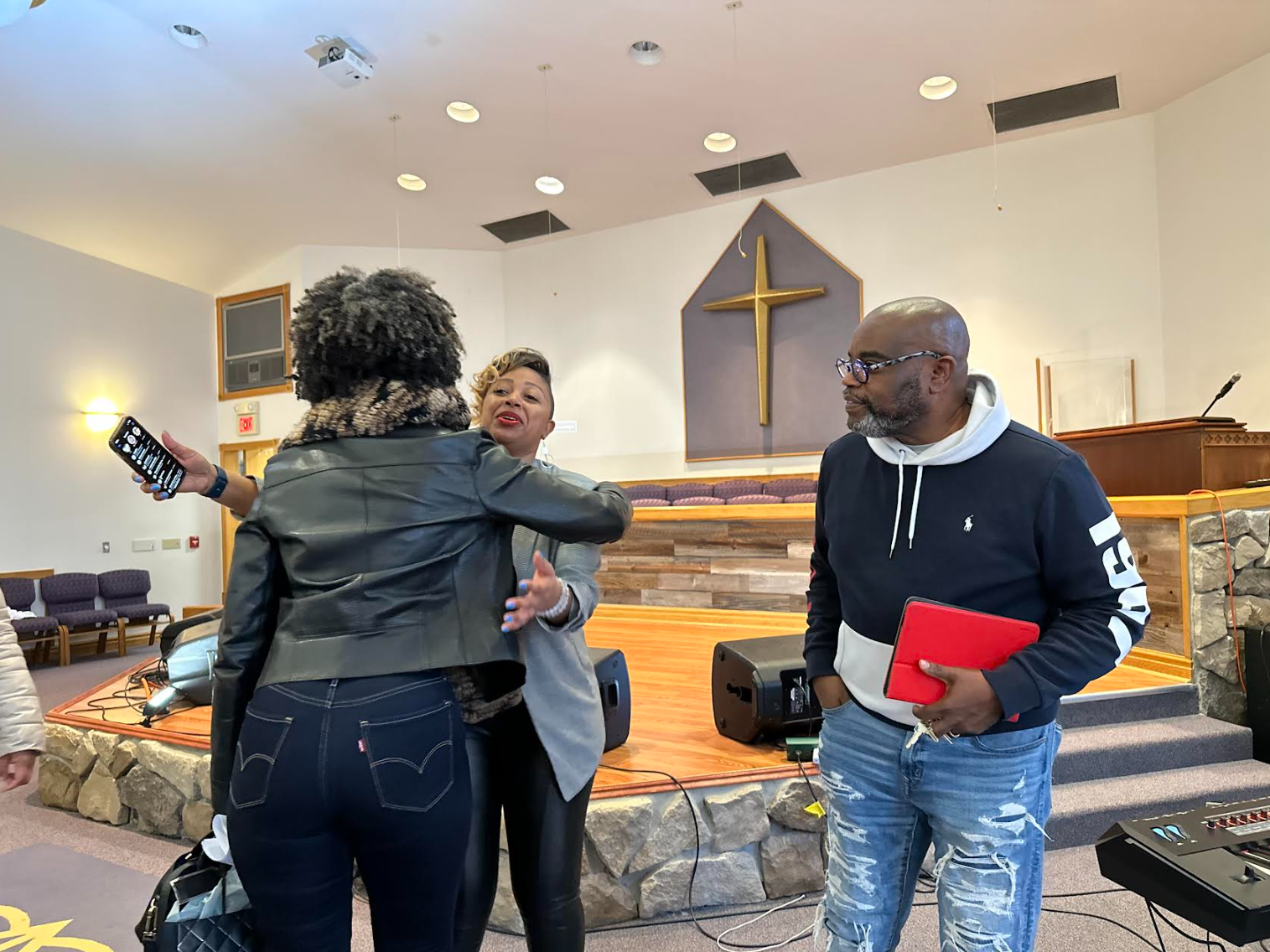 Three people in a church setting, with a woman hugging another woman near the altar, and a man standing nearby holding a tablet.
