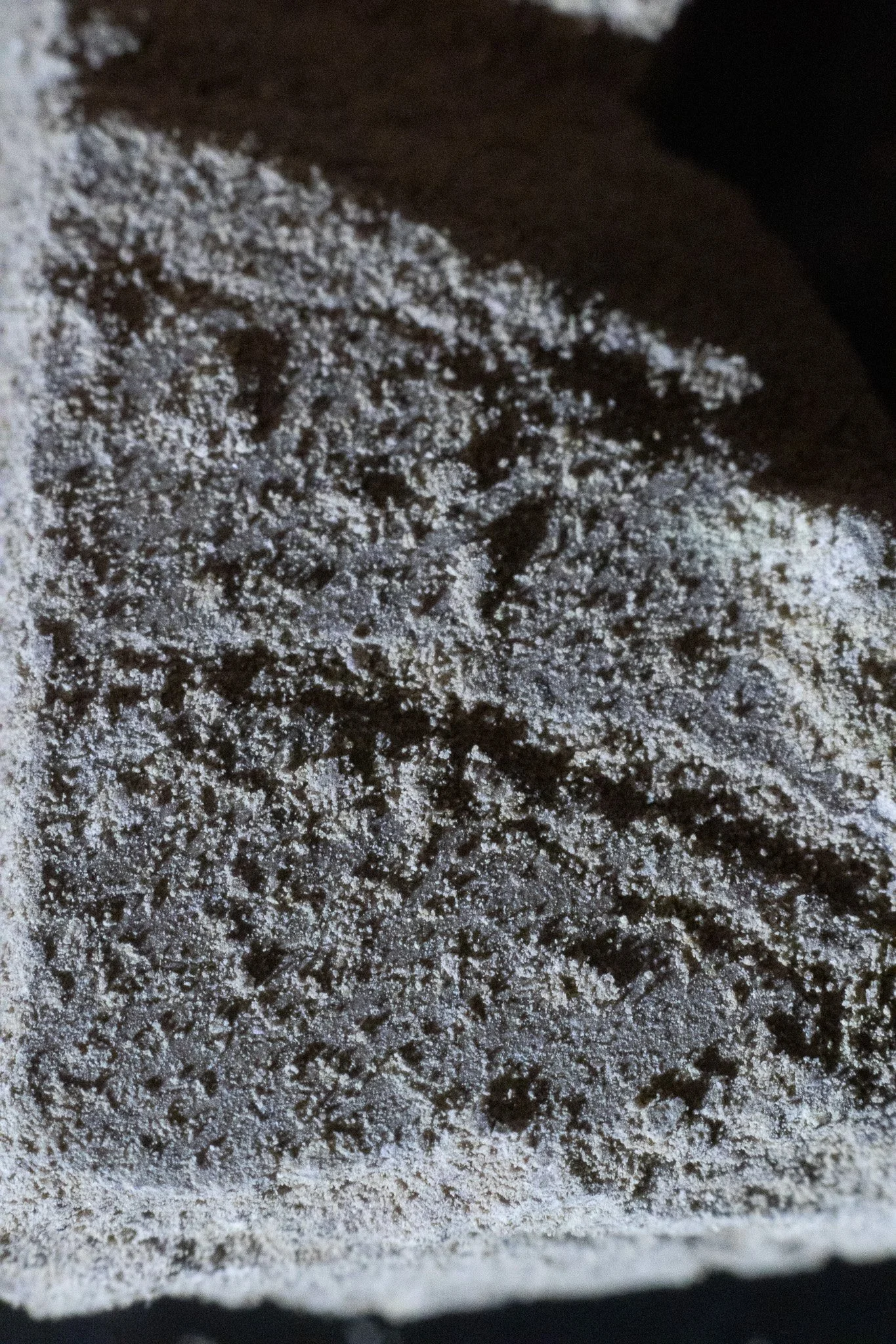 Close-up of a dark chocolate bar dusted with a white powder, possibly powdered sugar or flour, on a dark surface.