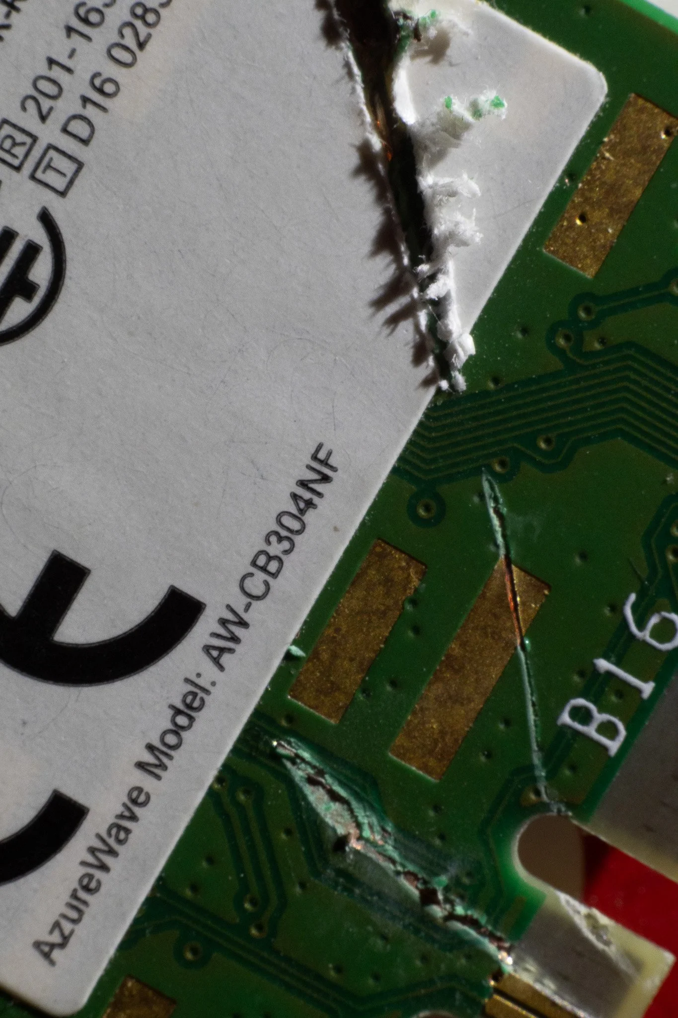 Close-up of a damaged printed circuit board with a partially torn white label showing model number, and burnt or schorched areas on the green circuitry.