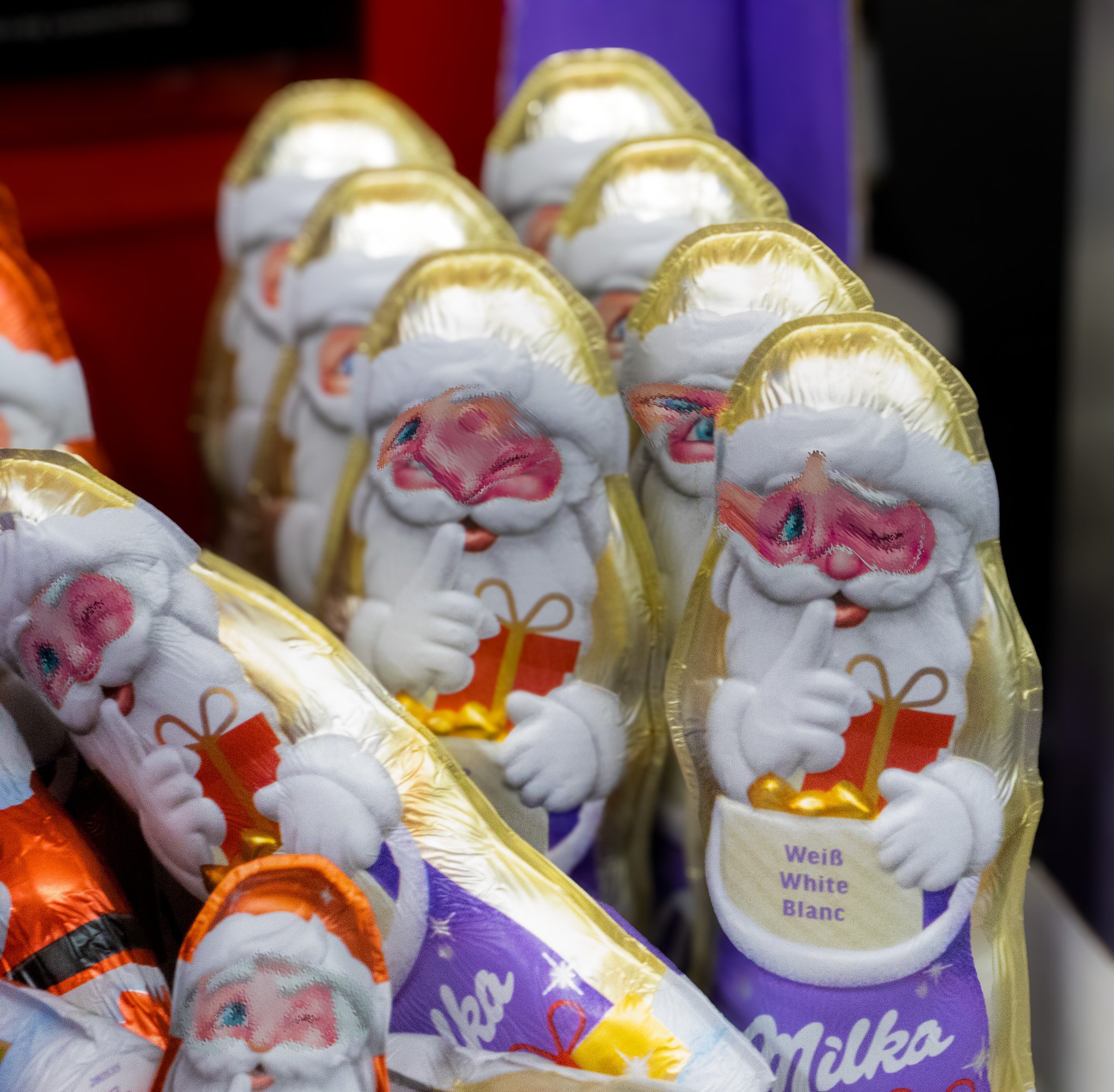 Multiple foil-wrapped chocolate Santa Claus figures with white beards, red noses, and smiling faces, each holding a gift, on a store shelf.