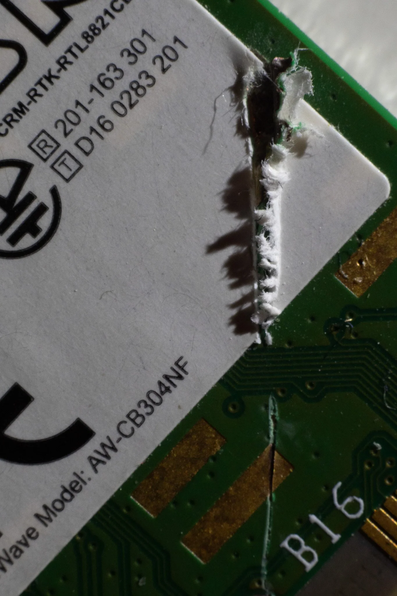 Close-up of a damaged circuit board with burnt area and melted components around a white label with model number AW-CB30ANE.