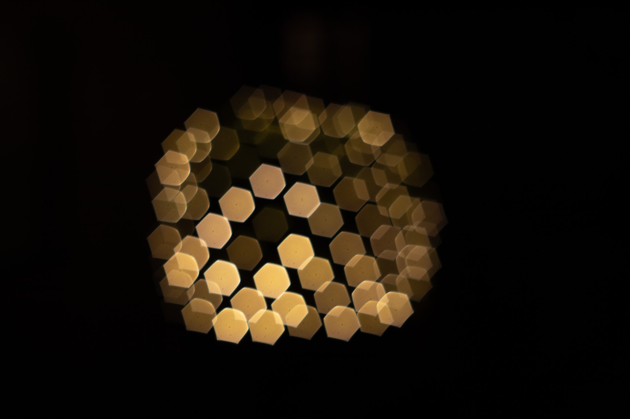 Out of focus image showing a grid of warm yellow-white lights arranged in a hexagonal pattern in a dark setting.