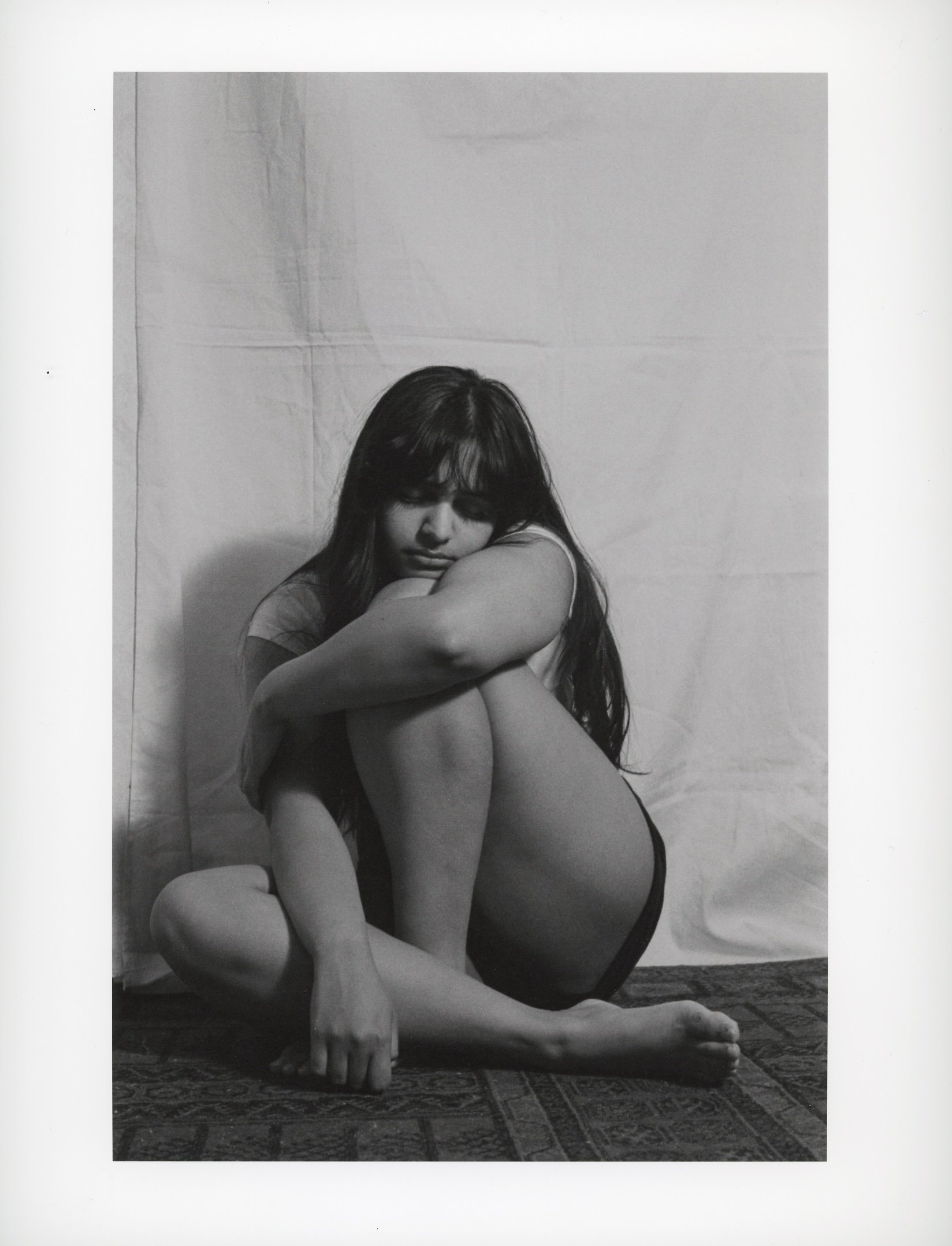 A black-and-white photograph of a young woman sitting on the floor with her arms wrapped around her knees, with her eyes closed and head resting on her arm. She has long, dark hair and is wearing a t-shirt and shorts.