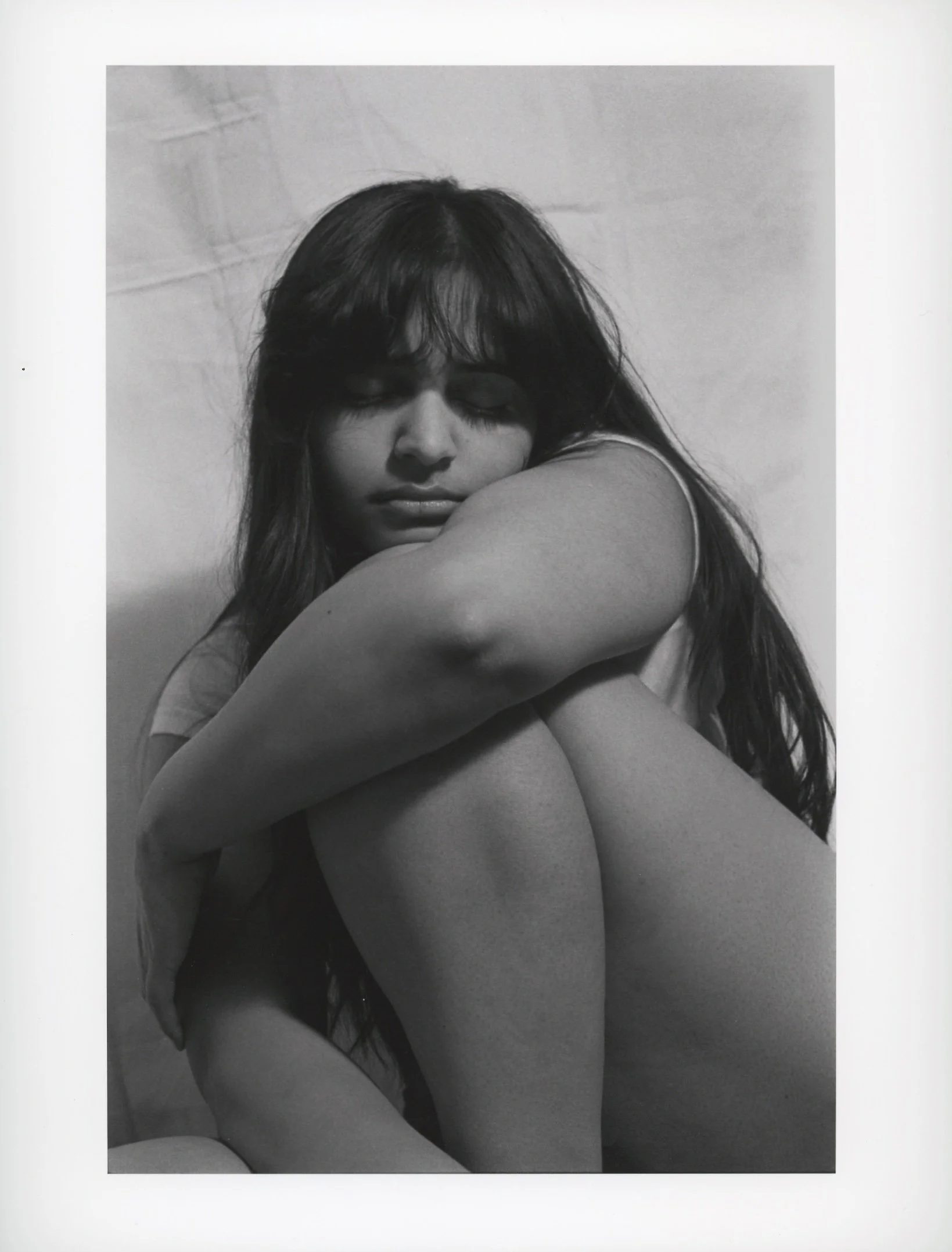 A black and white photograph of a woman with long hair sitting with her arms wrapped around her knees and eyes closed.