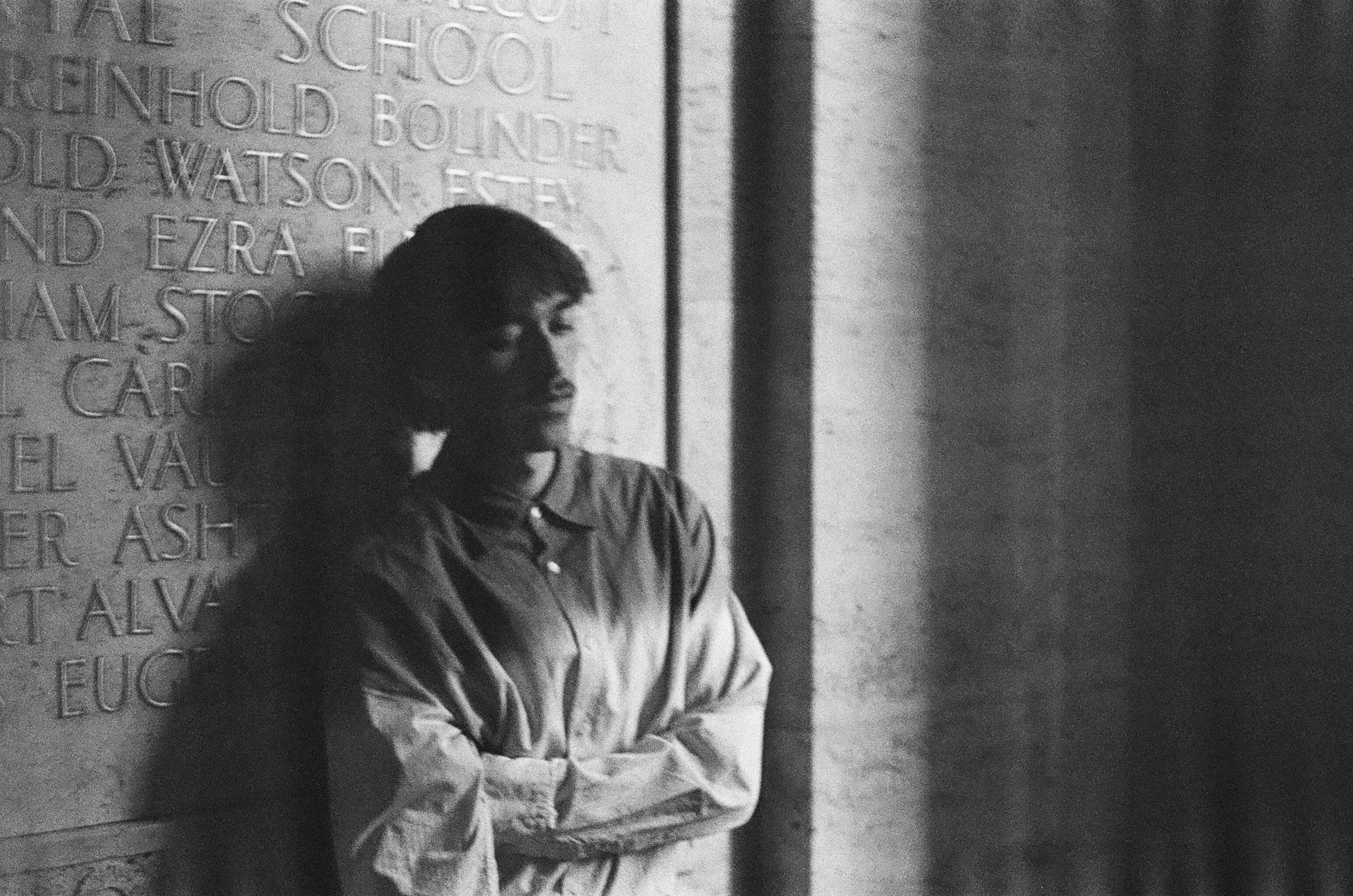 Black and white photo of a woman with her arms crossed, standing against a wall with engraved text.