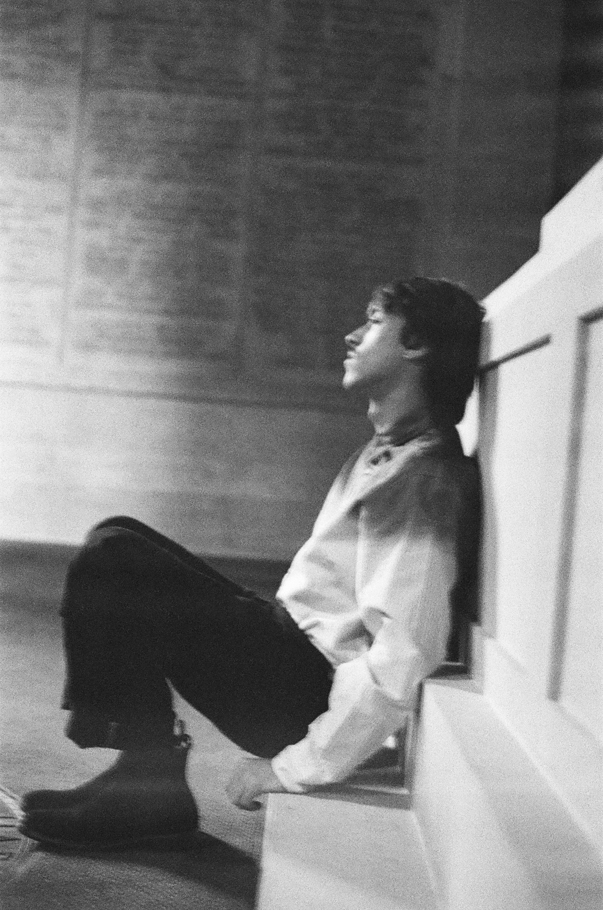 Black and white photo of a young person sitting on the floor with their back against a wall, looking to the side.
