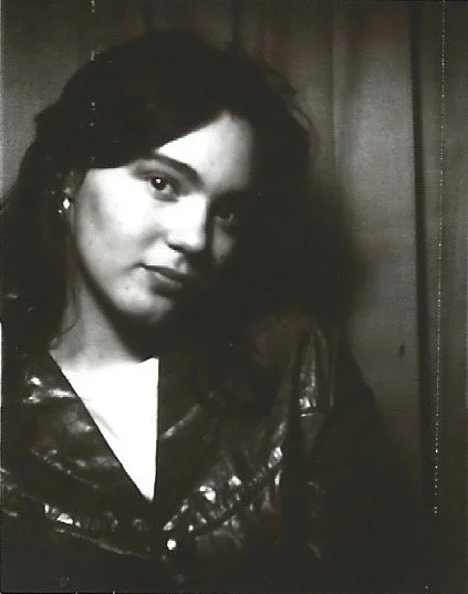 Black and white photo of a woman with dark hair, wearing a leather jacket, smiling slightly, in front of a curtain background.