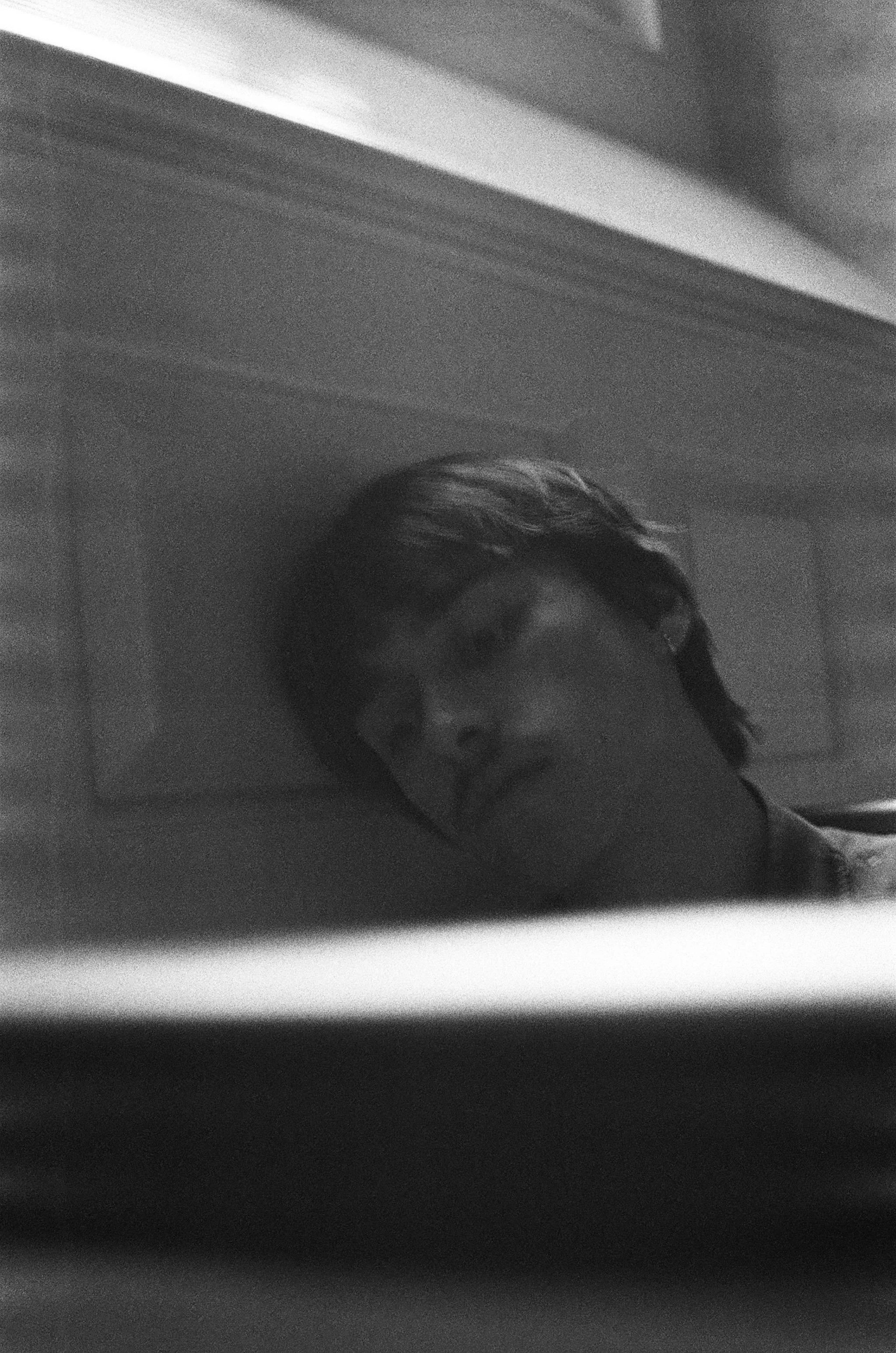 A black and white photo of a person with glasses, lying down with their head resting on a surface, looking at the camera.