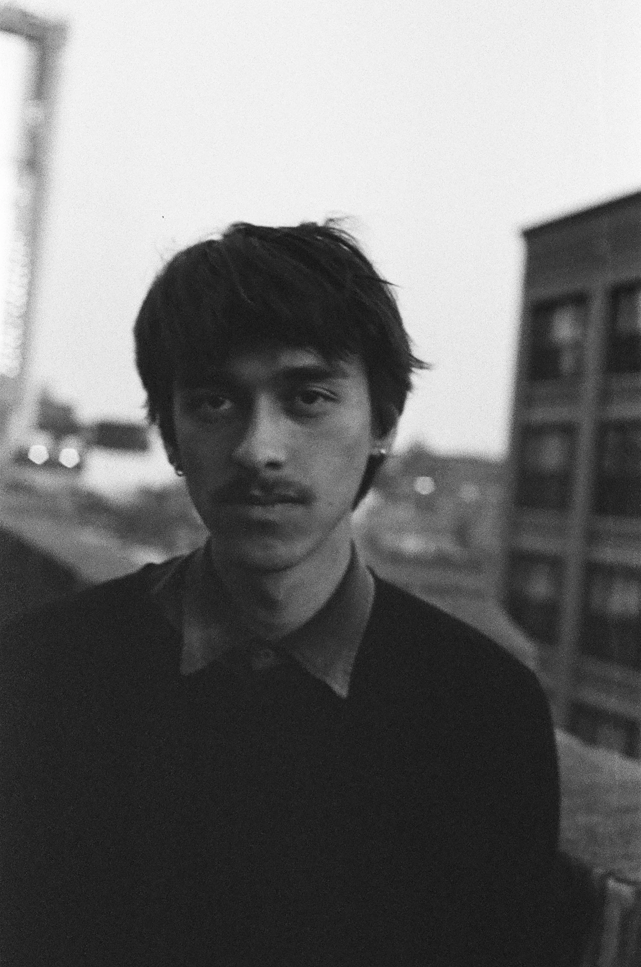 Black and white photograph of a young man with dark hair, standing outdoors on a city street during dusk or dawn, with buildings and a vehicle in the background.