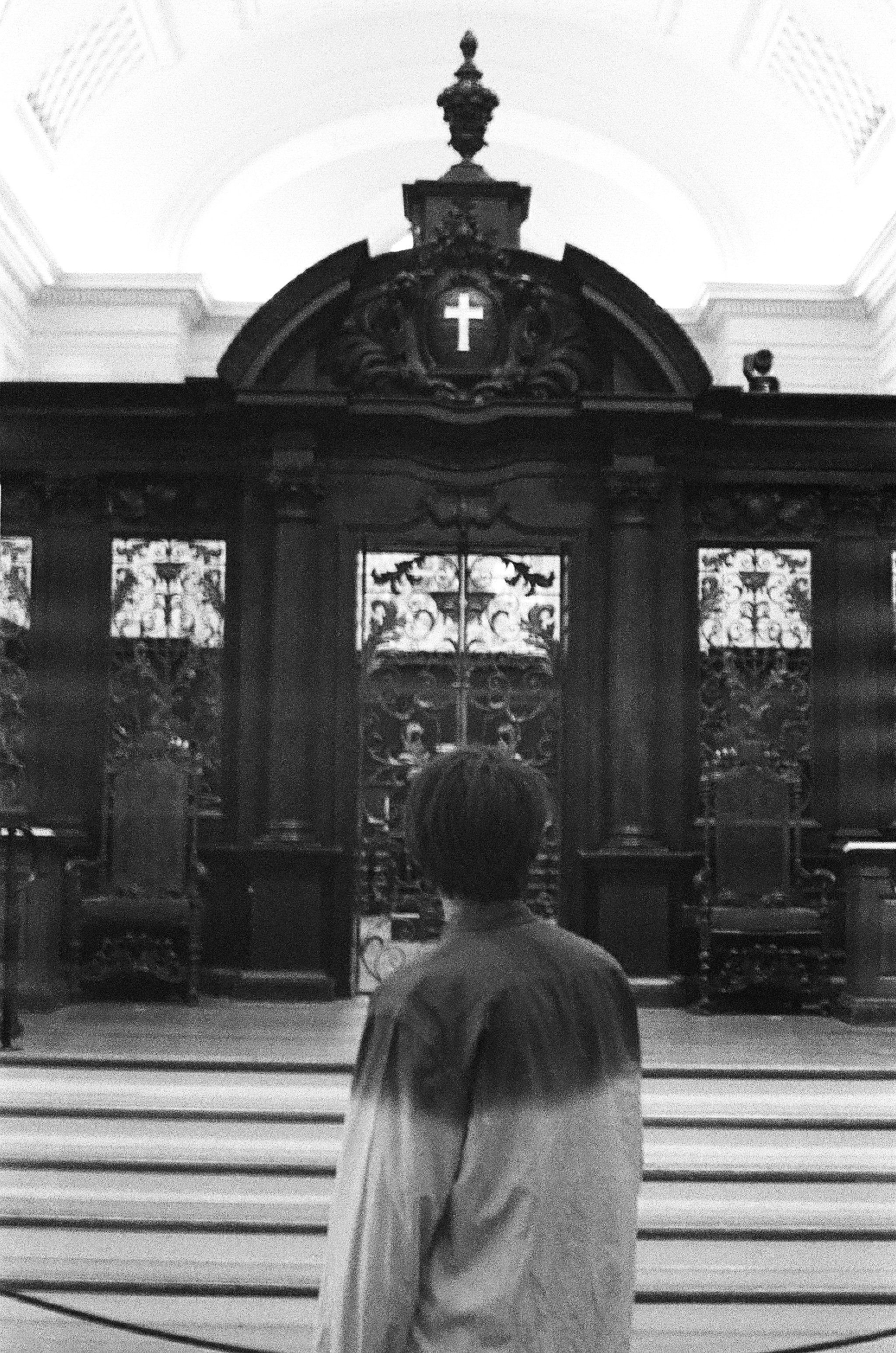 A person with short hair stands inside a church, facing an ornate wooden altar with intricate ironwork and a cross at the top.