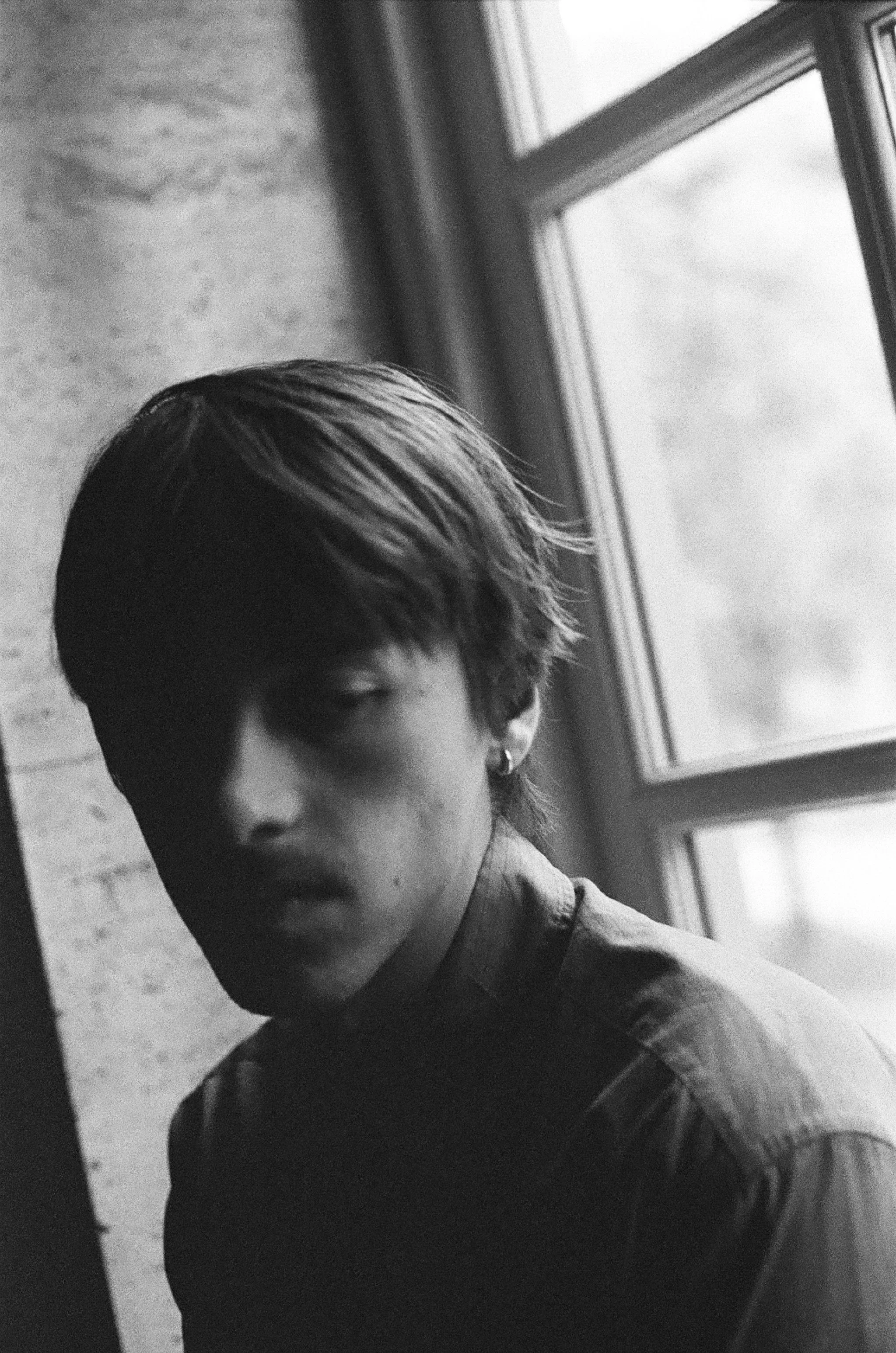 Black and white photo of a young person with short hair, wearing a shirt, looking at the camera with a serious expression, standing next to a window with light coming through.
