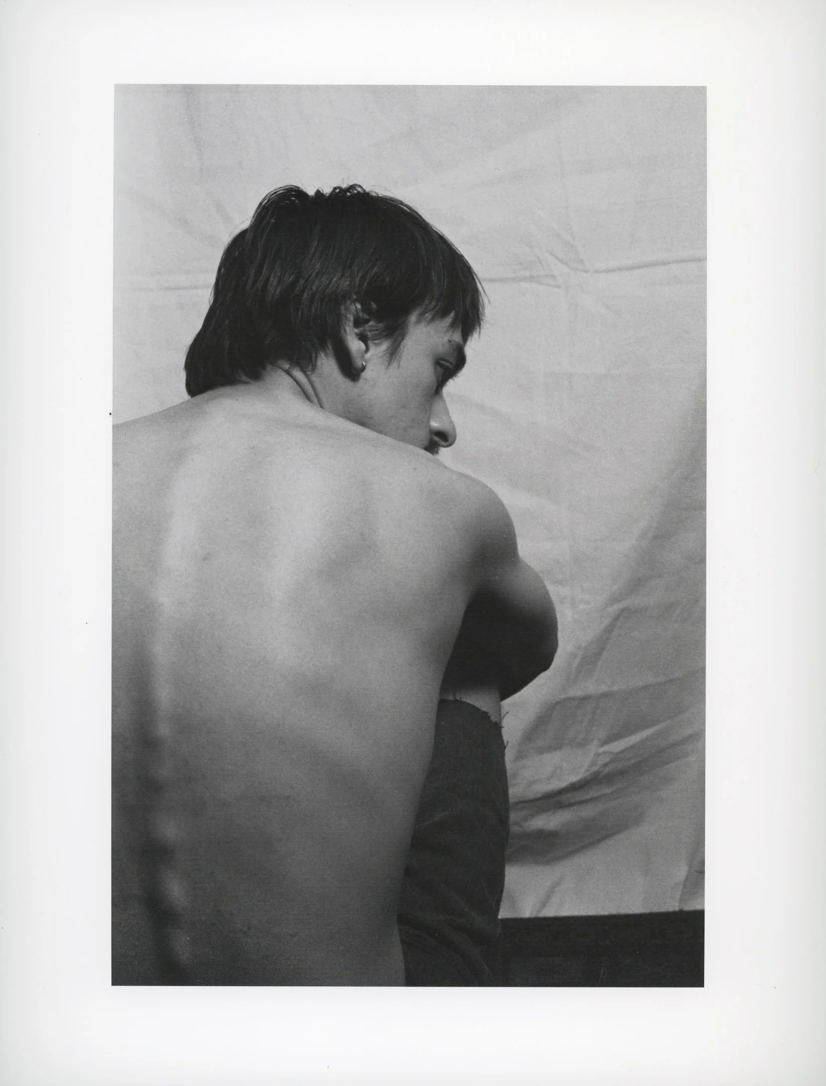 Black and white photograph of a shirtless person with short hair, viewed from the back, looking to the right, against a plain backdrop.