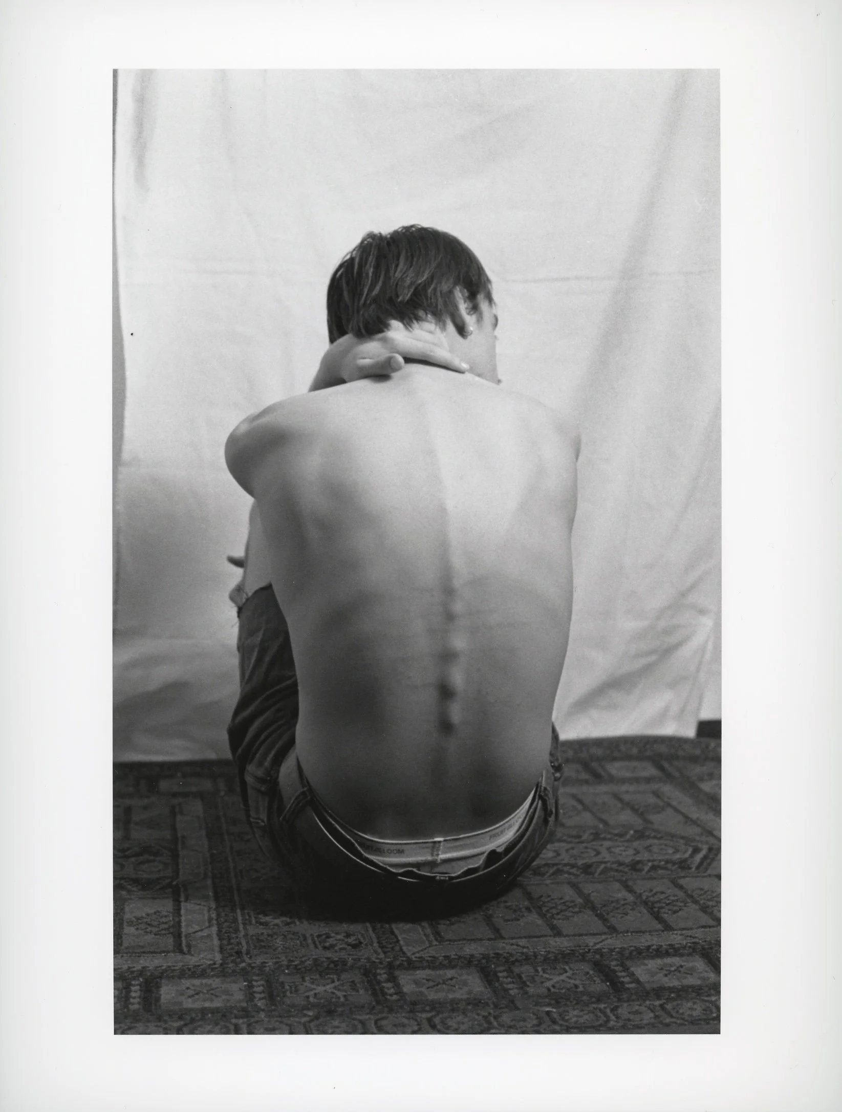 A black and white photo of a shirtless young man sitting on a patterned carpet with his back to the camera, embracing a person with their arms around his neck.