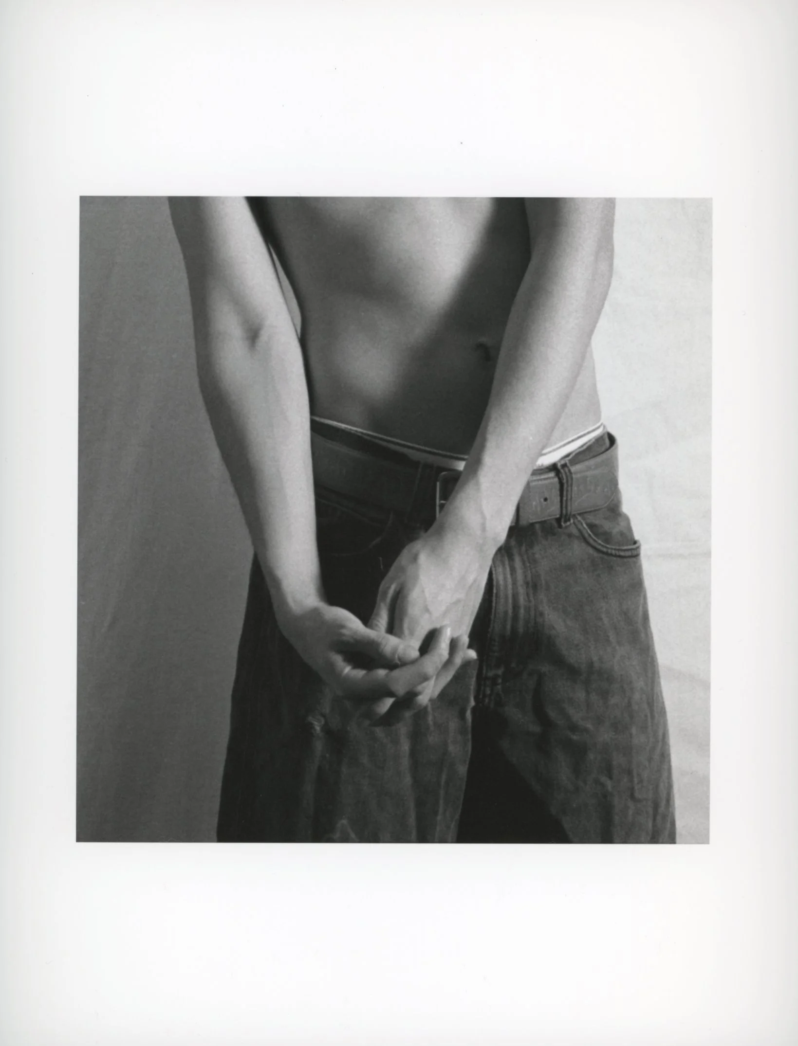 Black and white photo of a shirtless person with their hands clasped in front of them, wearing jeans and a belt.