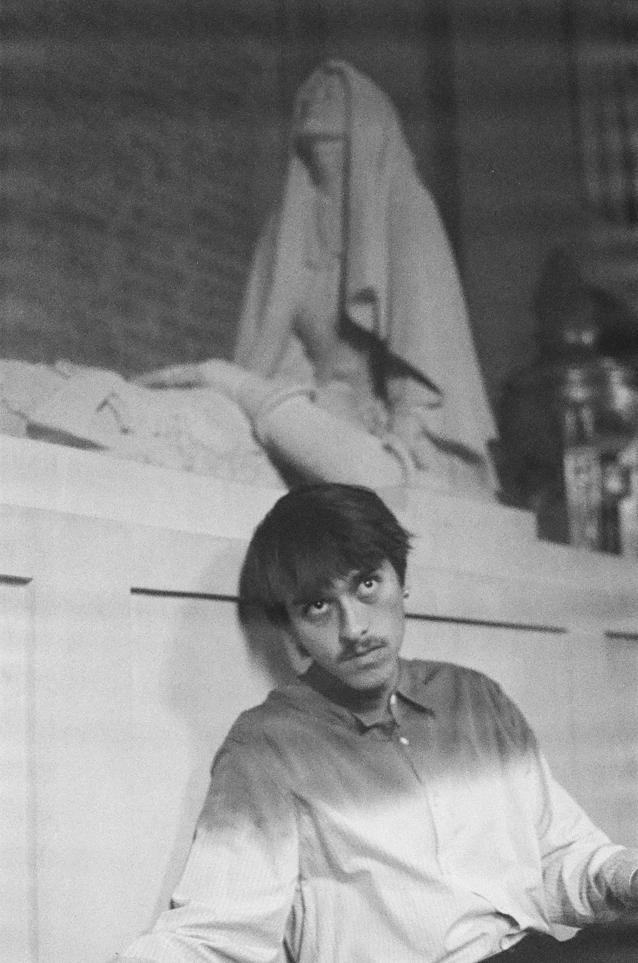 A young man with dark hair and a mustache sitting against a wall, looking up, with a woman sitting on a ledge in the background.