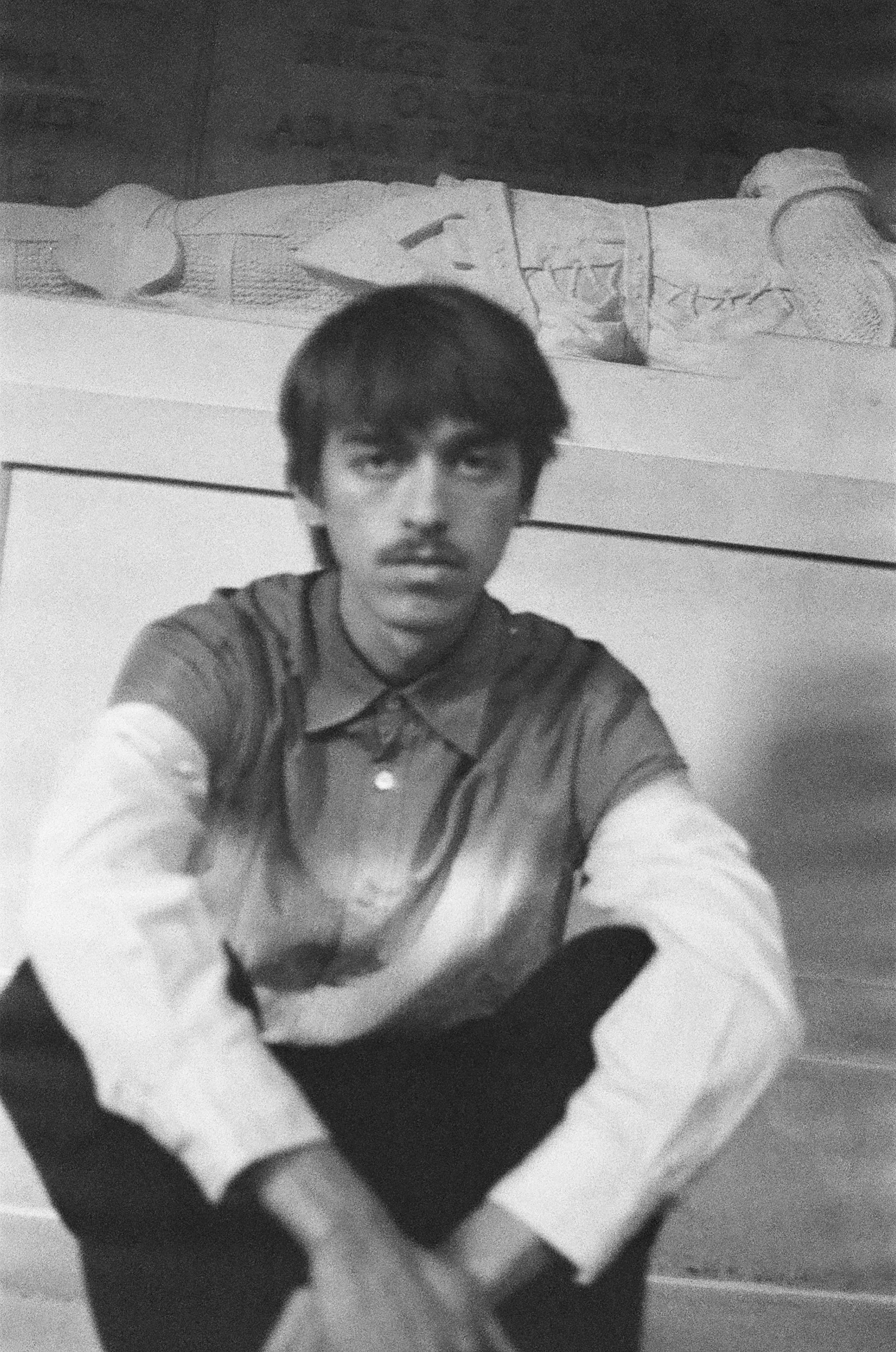 A young man with dark hair and mustache taking a selfie, sitting on the floor in front of a bed with a person lying on it. The photograph is black and white.